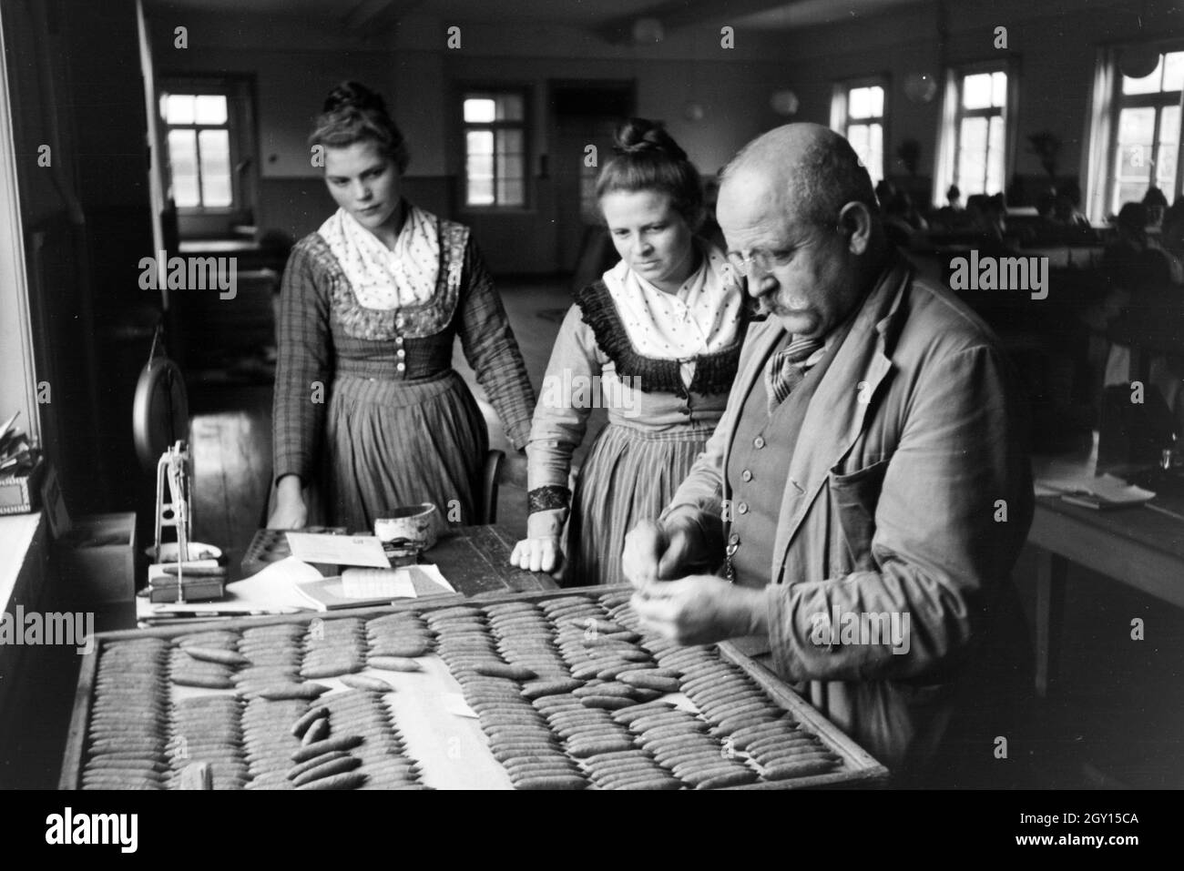 Der Besitzer einer Zigarrenfabrik bei Marburg erklärt-in Marburger Tracht gekleideten Mitarbeiterinnen sterben Qualitätskontrolle der fertigen handgerollten Zigarren, Deutschland 1930er Jahre. Der Besitzer eines Cigar Company in der Nähe von Marburg ist explaing die Qualitätskontrolle des fertigen handgerollte Zigarren Zigarre zu seinen weiblichen Rollen, tragen ein Gewand Marburg/Kostüm, Deutschland 1930. Stockfoto