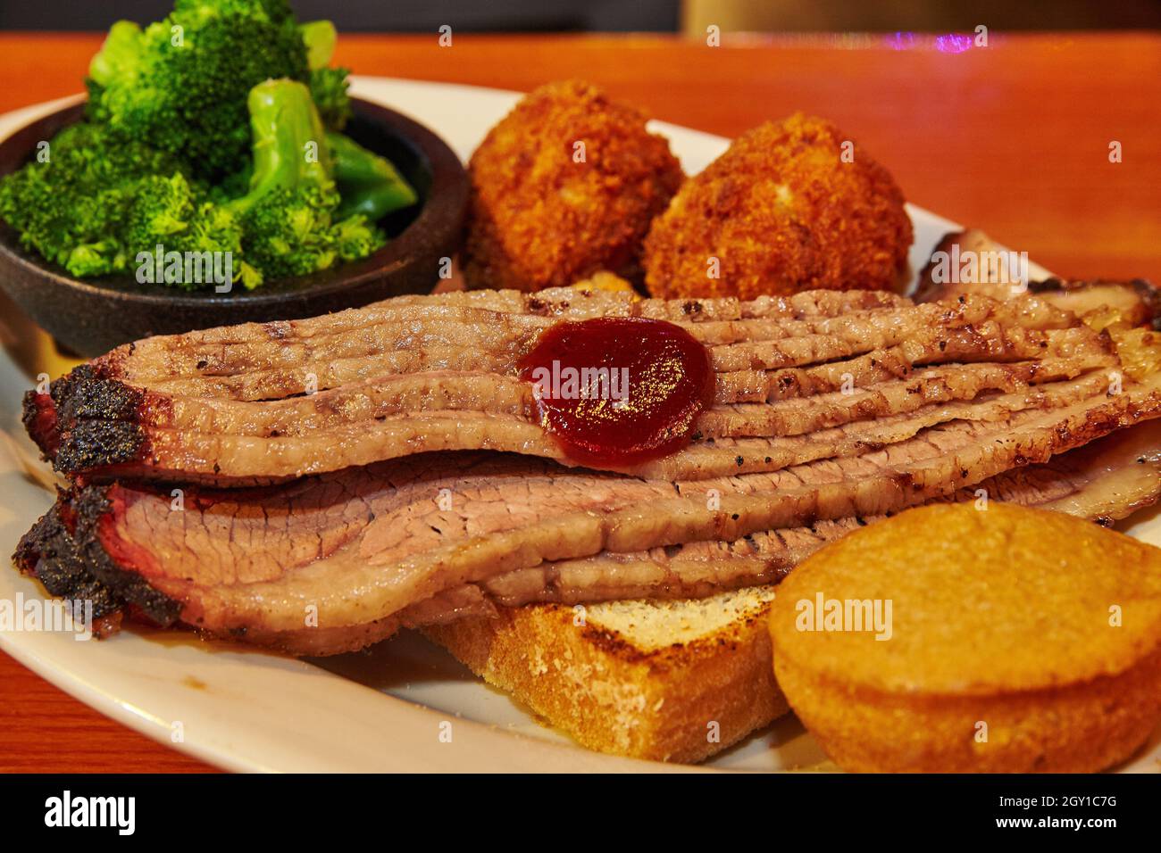 Scheiben Brisket mit Brot, Maismuffin, Hush Welpen, und Seite von Brokkoli auf einem weißen Teller Stockfoto