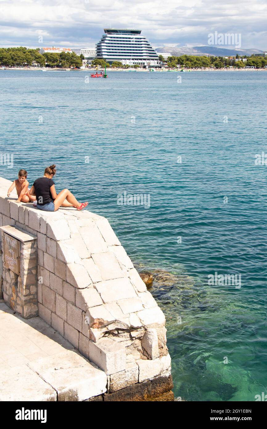 25. August 2021: Menschen, die auf einem Steinpier am Meer sitzen, und das moderne Hotelgebäude Olympia Sky in der Ferne, aus dem hohen Winkel Stockfoto