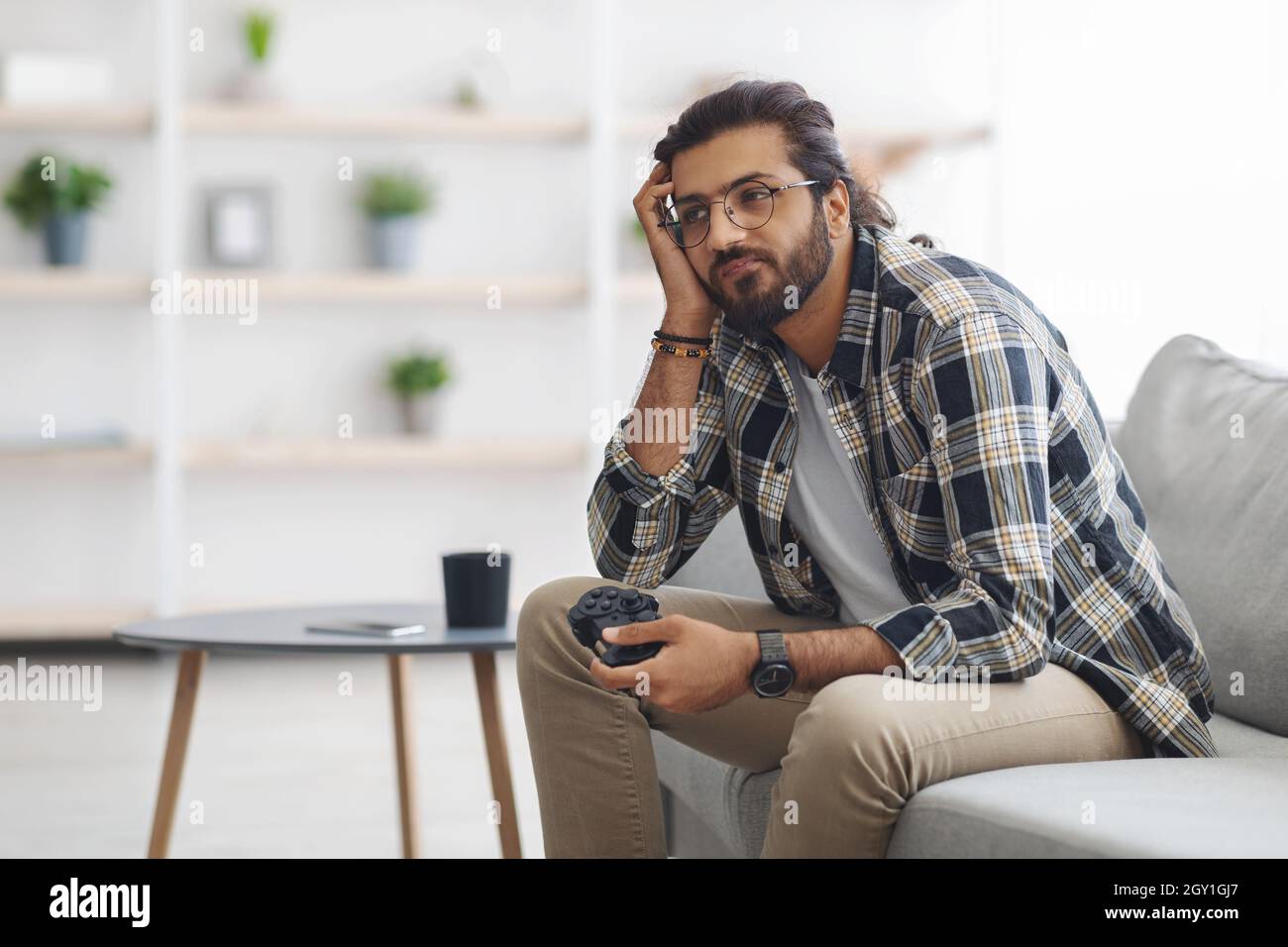 Gelangweilter indischer Kerl, der mit Joystick auf der Couch sitzt Stockfoto