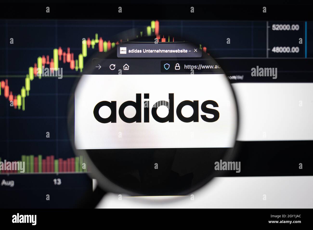 Adidas Firmenlogo auf einer Website mit verschwommenen Börsenentwicklungen im Hintergrund, die auf einem Computerbildschirm durch eine Lupe gesehen werden Stockfoto