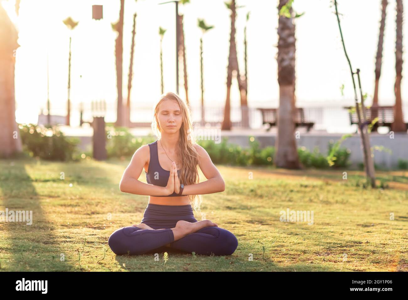 Junges Mädchen, das in Lotus-Pose im Park in der Nähe der Strandlinie bei Sonnenuntergang Sonnenaufgang sitzt. Schönes Mädchen praktiziert Yoga während Sommerurlaub Meditation SE Stockfoto