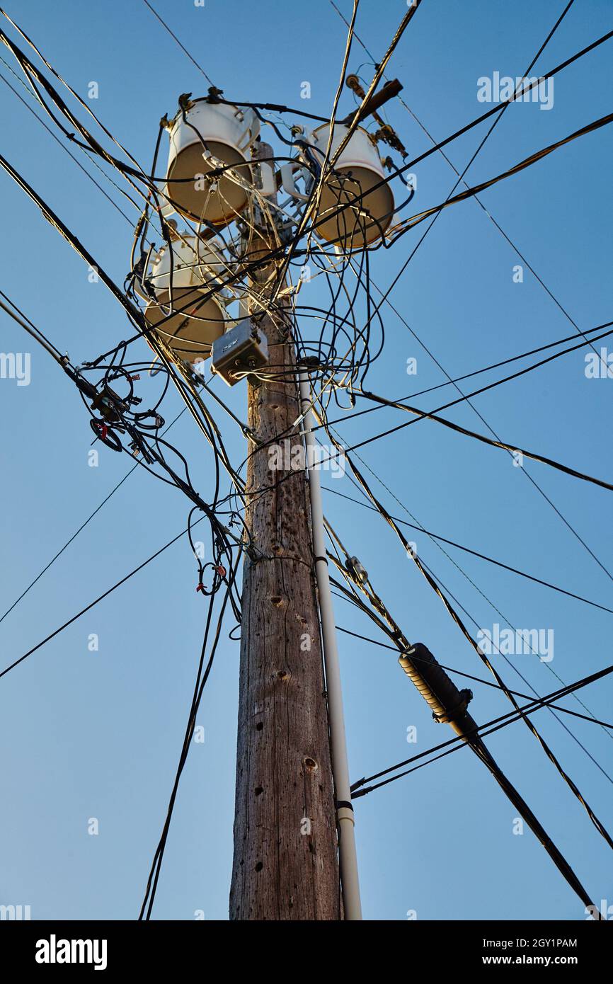 Hölzerner Telefonmast mit drei Fässern oben und vielen konvergierenden Telefonleitungen an einem klaren blauen Himmel Stockfoto