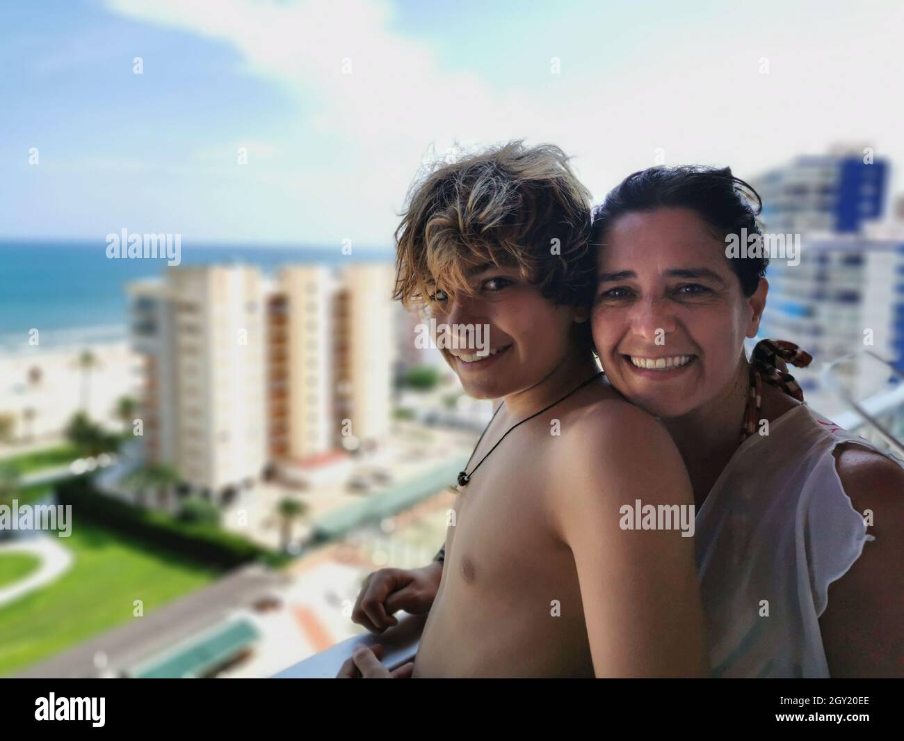 Die fröhliche hispanische Mutter und ihr Sohn umarmen sich auf dem Balkon mit Blick auf die Stadt und das Meer Stockfoto