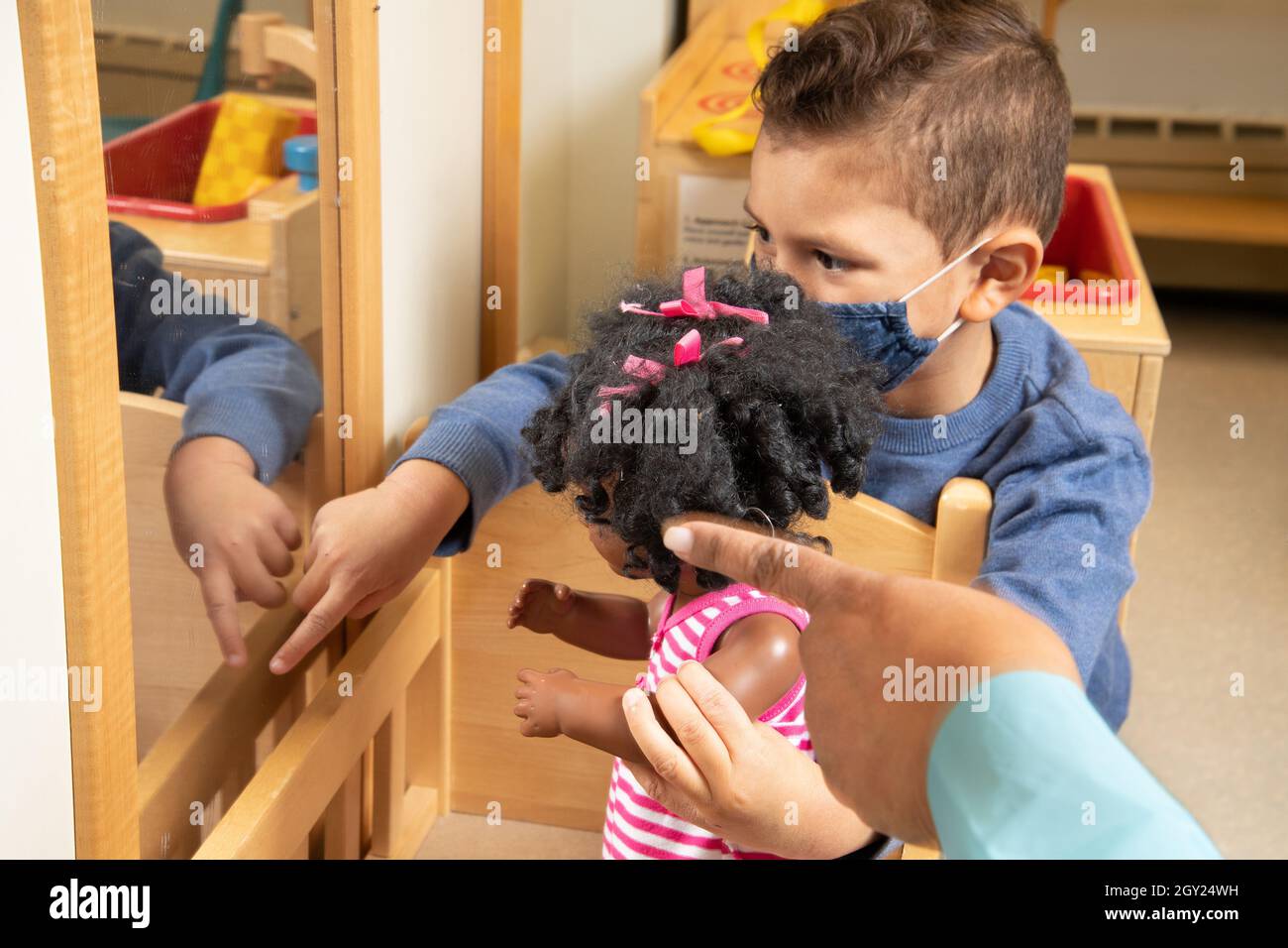 Bildung Preschool Junge im Alter von 3-4 Jahren im Familienbereich hält Puppe zum Spiegel und zeigt auf Reflexion, während die Hand des Lehrers von der Seite nach innen zeigt und das fa trägt Stockfoto