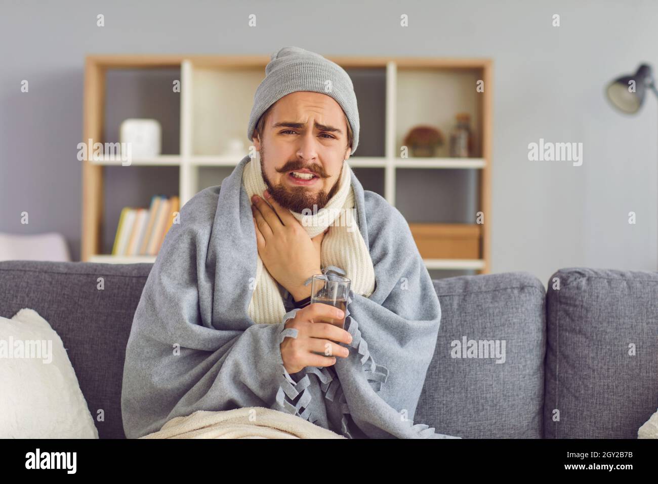 Junger Mann in warmen Kleidern und Decken sitzt auf dem Sofa mit einem Glas Wasser, berührt den Hals und fühlt sich schlecht Stockfoto
