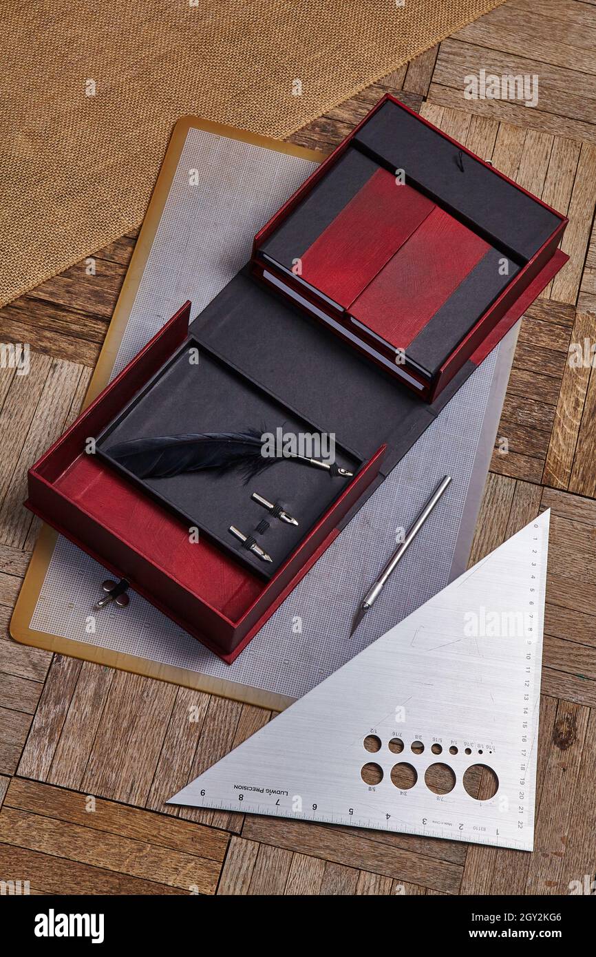 Rote und schwarze offene Box mit einer Federkiel liegt neben einem Gebrauchsmesser und einer dreieckigen geraden Kante Stockfoto