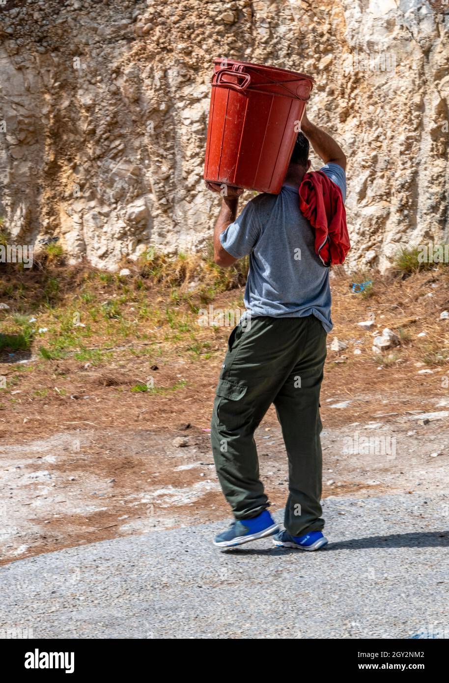 Arbeiter, der einen schweren roten Mülleimer auf der Schulter trug, Mann mit Mülleimer auf der Schulter, der Müll leerte. Mann, der schwere Last auf der Schulter trug. Stockfoto