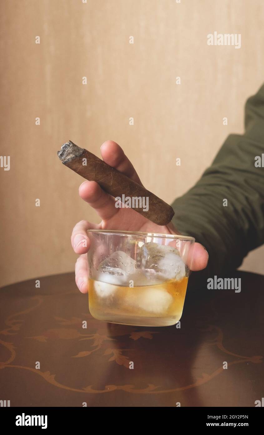 Ein Mann in einem grünen Hemd hält eine kubanische Zigarre und ein Glas Whiskey auf Eis. Nahaufnahme einer rauchenden Zigarre. Stockfoto