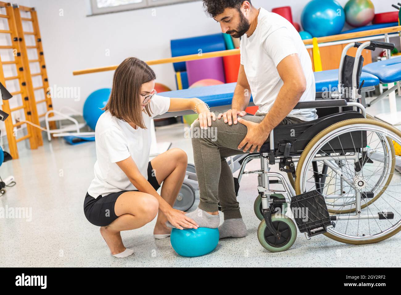 Pphysiotherapeut und männlicher Patient, der während der Rehabilitationsbehandlung im Rollstuhl sitzt - drücken und lösen Sie einen Fuß auf einen kleinen Übungsball Stockfoto
