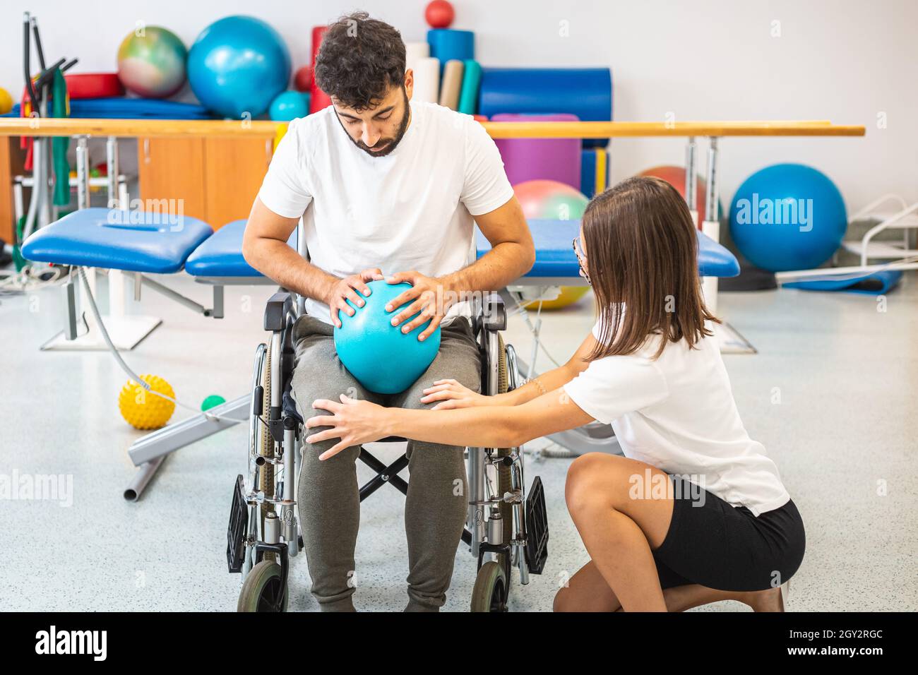 Behinderter Mann, der in einem Rollstuhl sitzt und eine Übung mit einem kleineren Pilates-Ball im physiotherapeutischen Rehabilitationszentrum macht Stockfoto