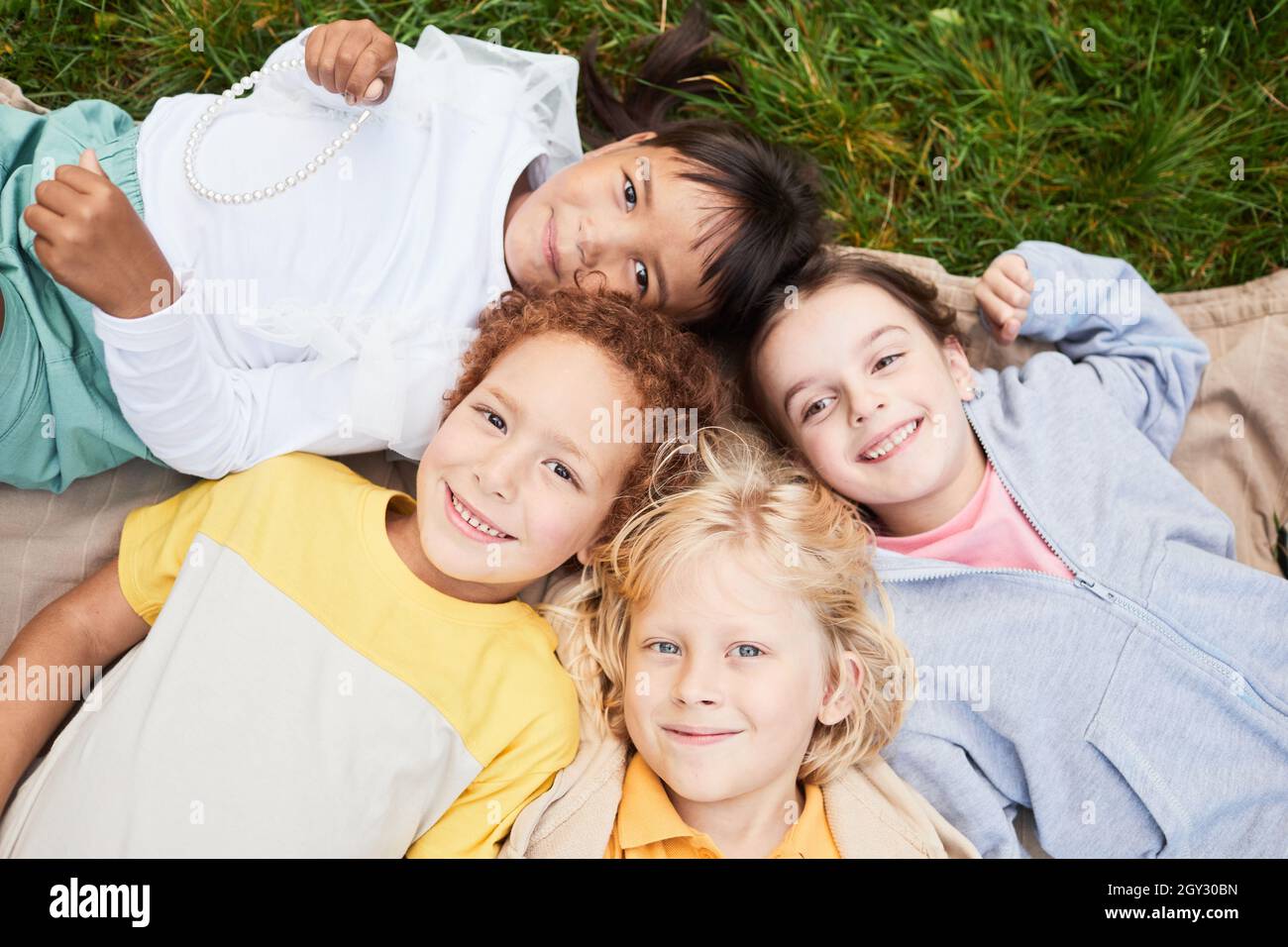 Draufsicht Porträt einer vielfältigen Gruppe von Kindern, die im Park auf Gras liegen und lächelnd auf die Kamera blicken Stockfoto