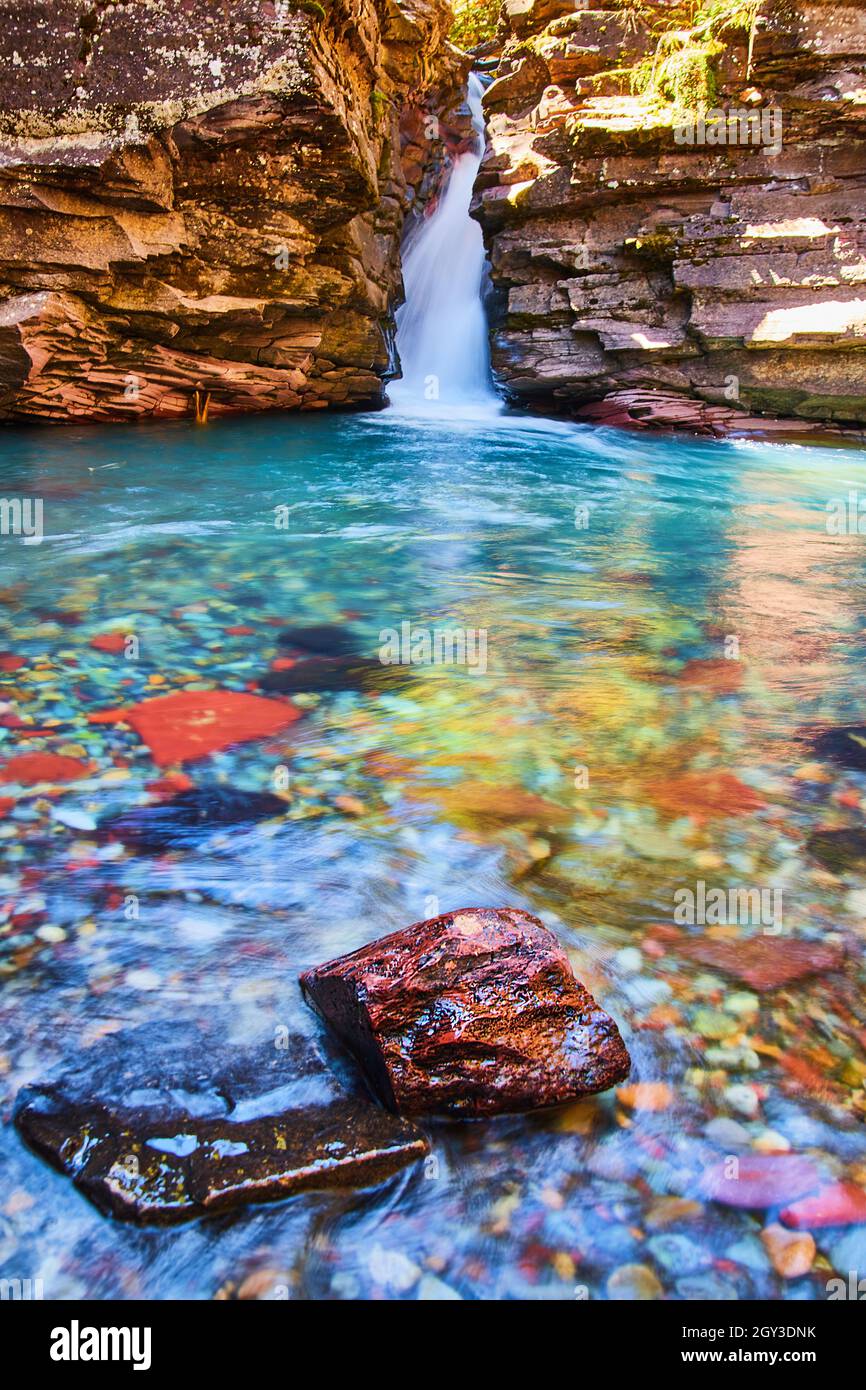Farbenfrohe Flussfelsen im Fokus mit großem Wasserfall und Klippen am unteren Rand der Schlucht Stockfoto