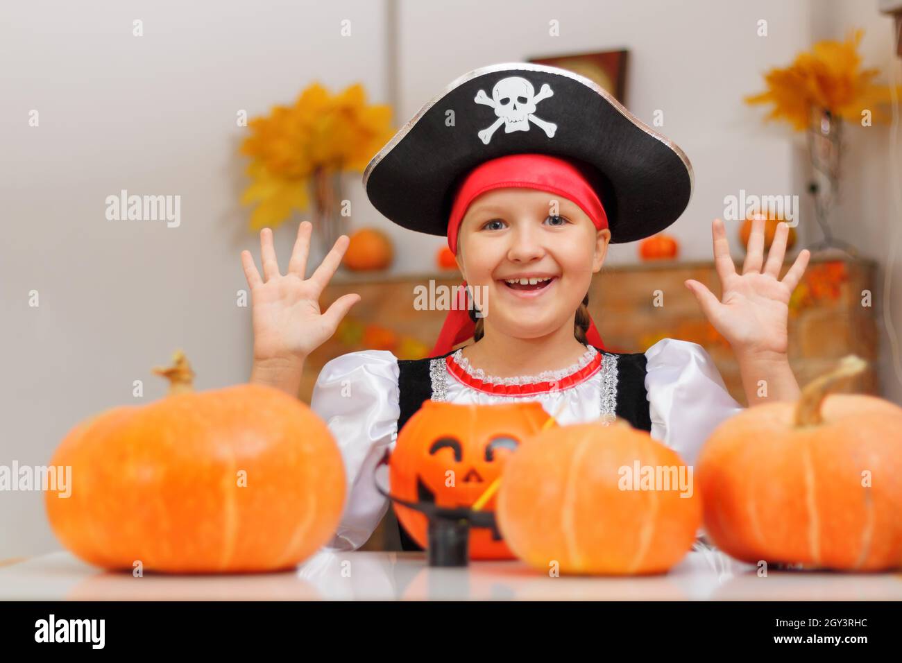 Halloween zu Hause feiern. Ein fröhliches kleines Mädchen in einem Piratenkostüm sitzt mit Kürbissen und einem Eimer Jack Laterne an einem Tisch. Das Kind zeigt Stockfoto