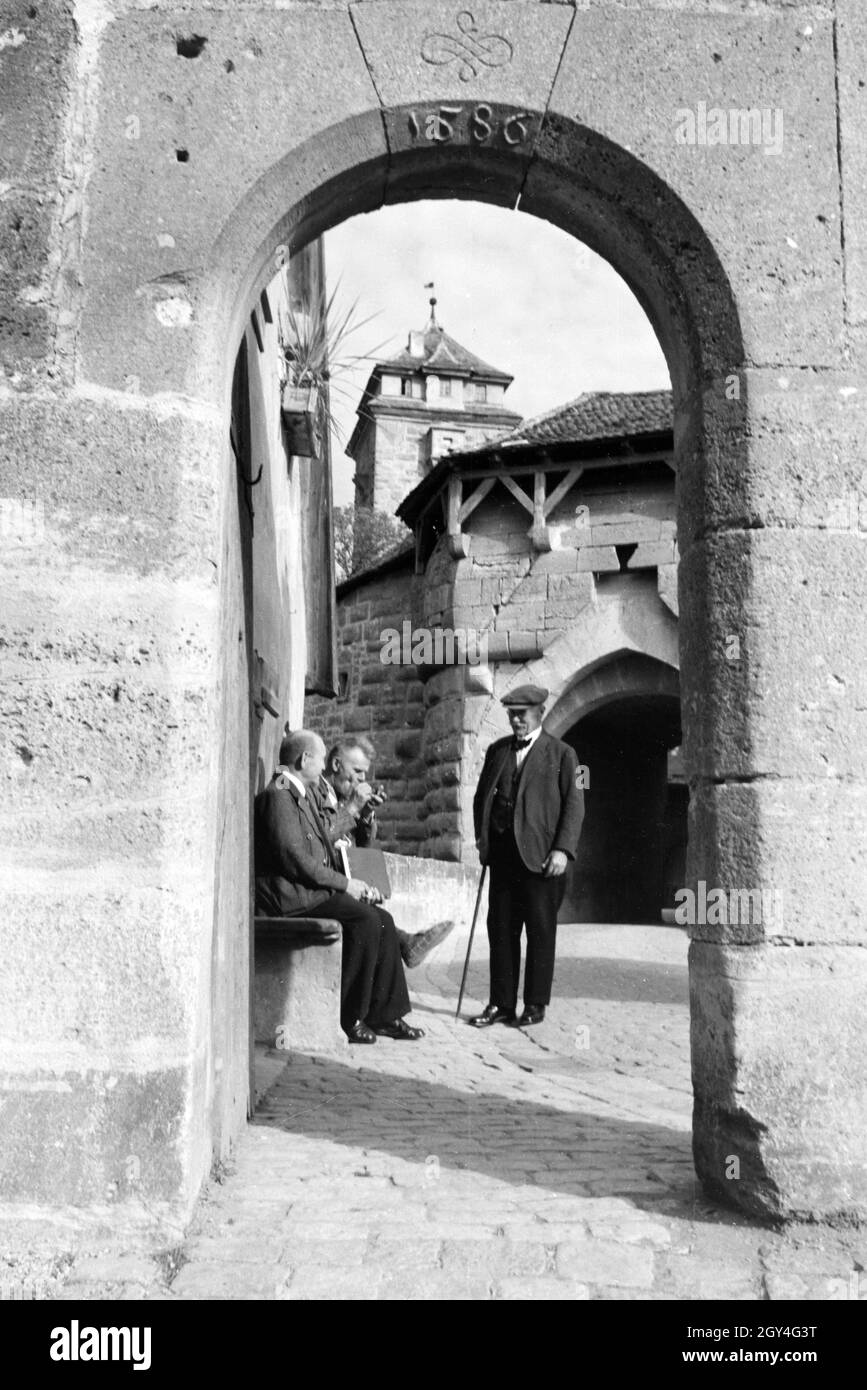 Drei ältere Herren plauschen auf einer Bank vor einem der zahlreichen historischen Torbögen in Rothenburg o.d. Tauber, Deutschland 1930er Jahre. Drei ältere Männer chitchating beim Sitzen auf einer Bank vor einem der vielen historischen Arkaden in Rothenburg o.d. Tauber, Deutschland 1930. Stockfoto