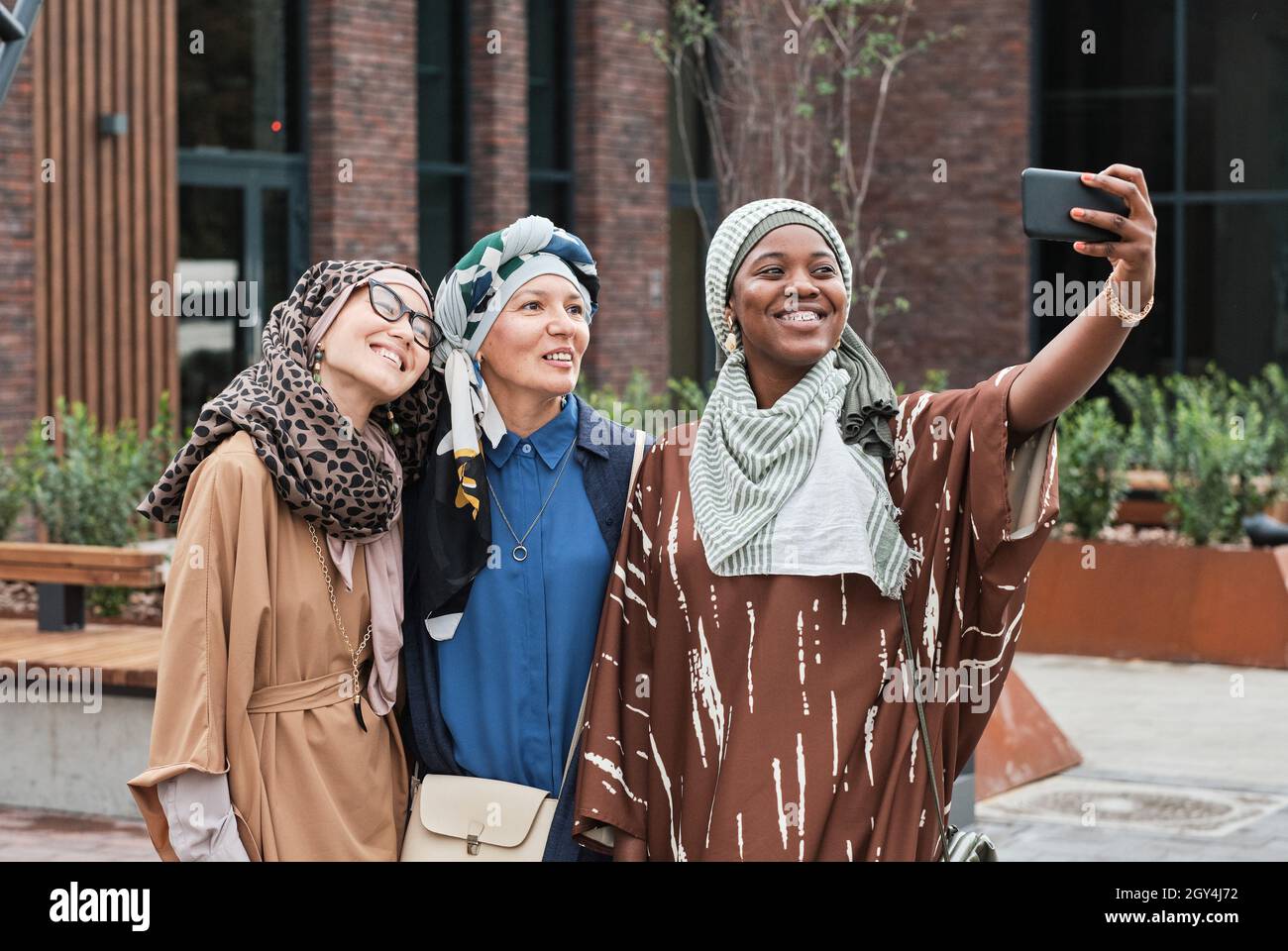 Drei elegante Frauen in muslimischer Kleidung lächeln der Kamera des Mobiltelefons zu, während sie im Freien Selfie-Portraits machen Stockfoto