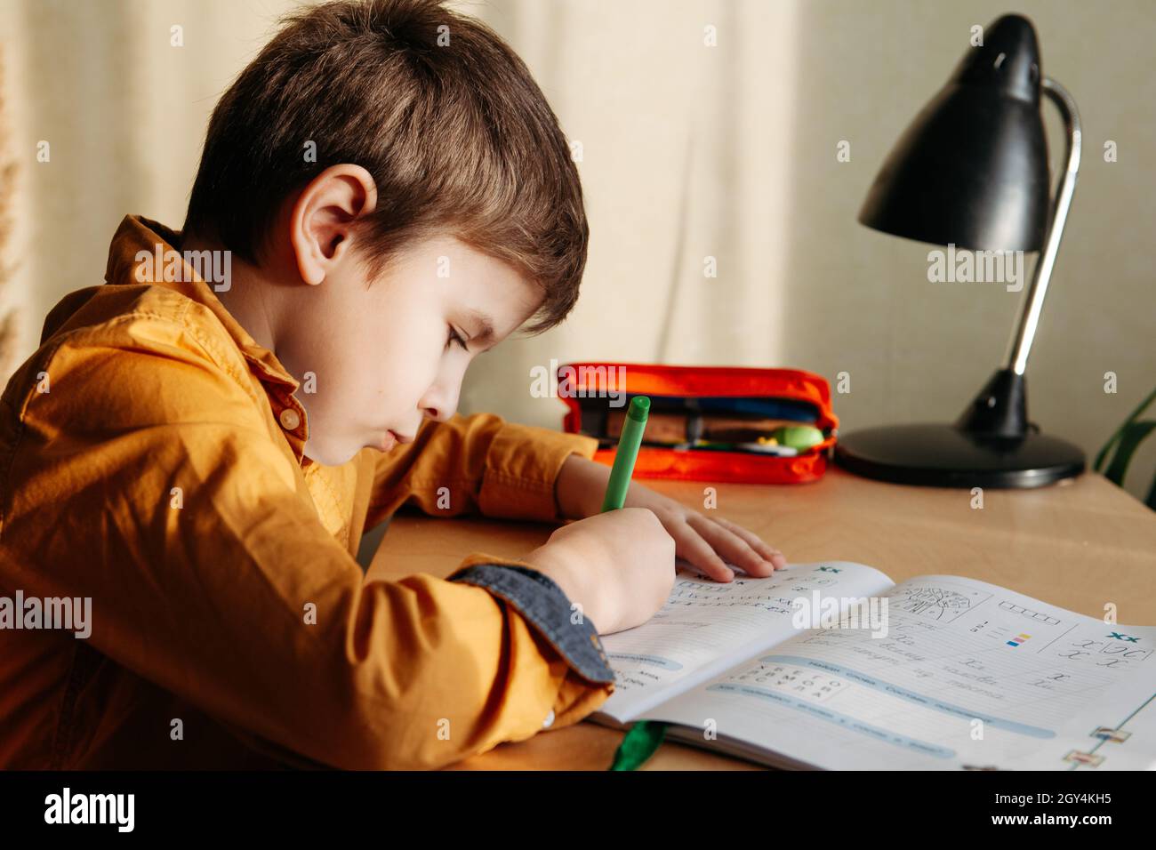 Niedliches 7-jähriges Kind, das seine Hausaufgaben am Schreibtisch macht. Junge schreibt im Notizbuch. Seitenansicht Stockfoto