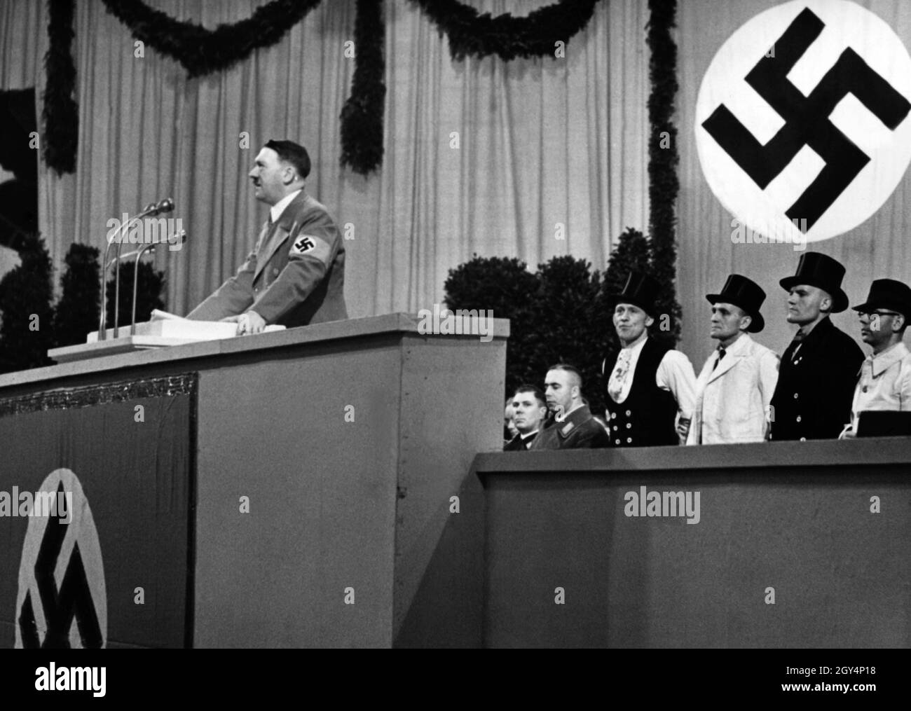 Hitler hält eine Rede im Sportpalast, um die Fertigstellung der neuen Reichskanzlei zu markieren. Hinter ihm stehen Vorarbeiter in Gildenkleid, die am Bau beteiligt waren. [Automatisierte Übersetzung] Stockfoto