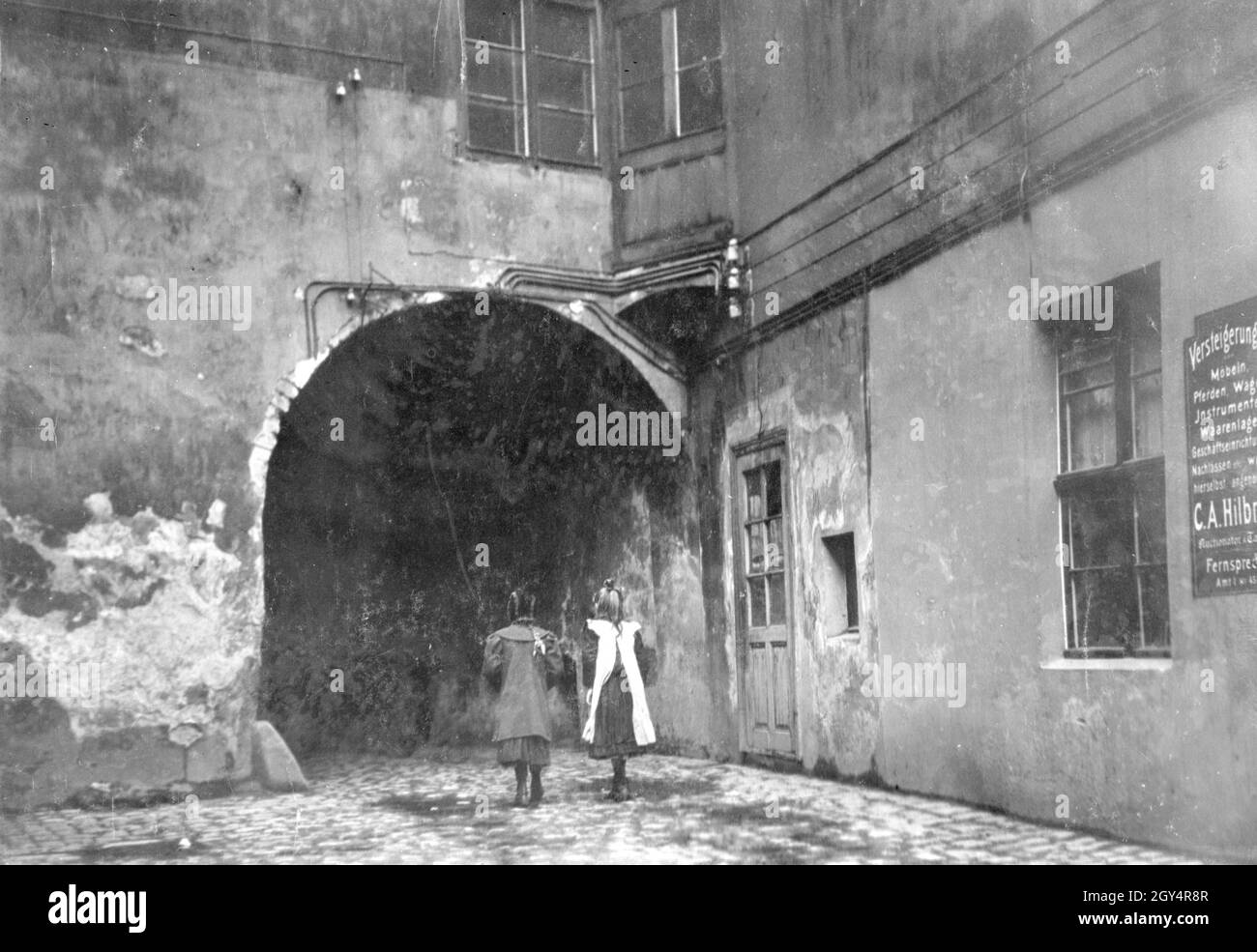 Zwei Mädchen gehen 1907 durch einen Torbogen in der Gasse am Krögel in Berlin-Mitte. Ein Brett auf der rechten Seite lautet: ''Auktionen von Möbeln, Pferden, Wagen, Instrumenten, Warenbestand, Geschäftsausstattung, Güter usw. werden hier akzeptiert. C. A. Hilbrecht.“ [Automatisierte Übersetzung]' Stockfoto