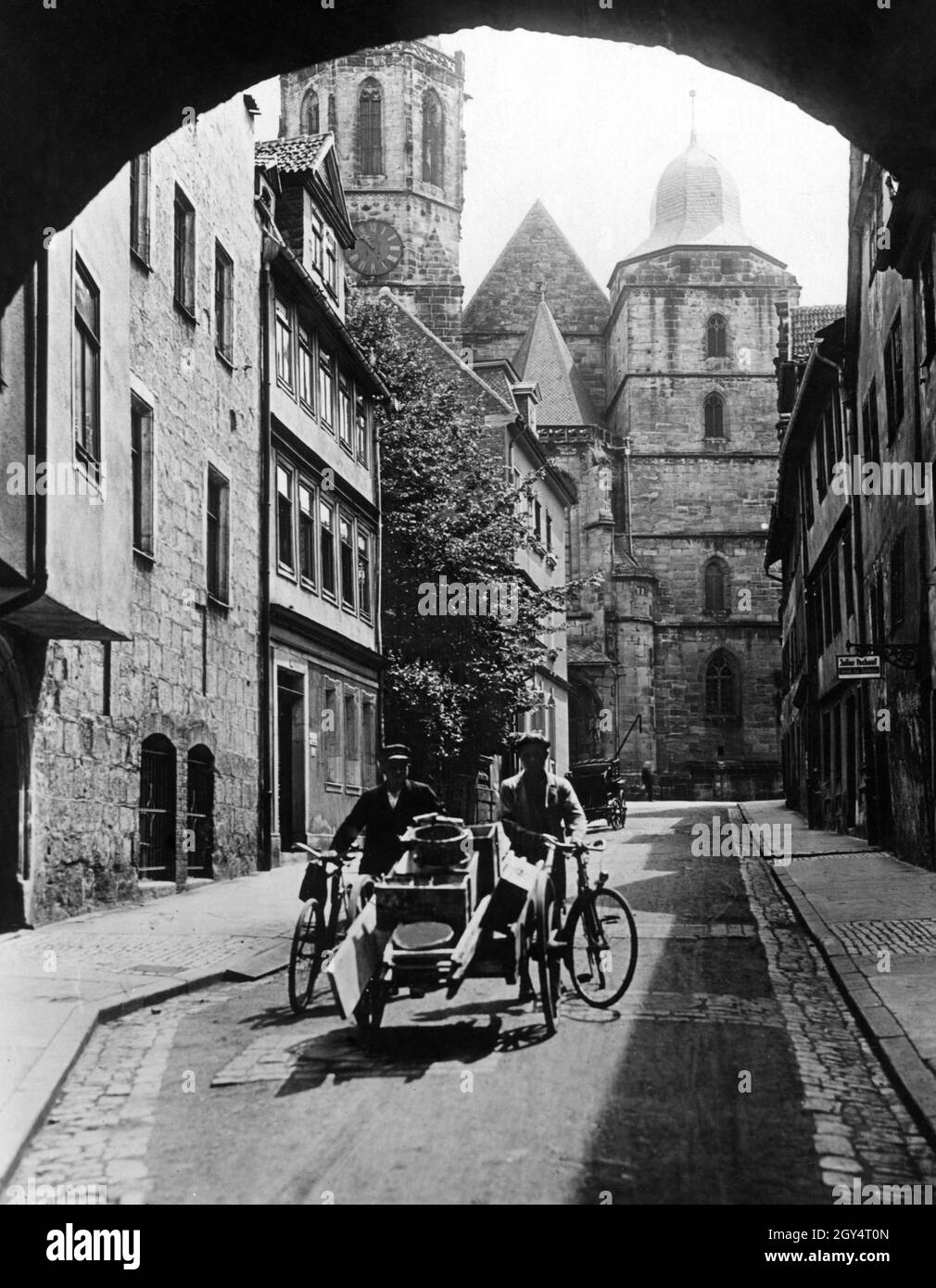 Der Durchgang unter dem Münzmeisterhaus in Coburg führt in die Neugasse und bietet einen Blick auf die Stadtkirche St. Moriz mit ihrem unvollendeten Südturm. Zwei Arbeiter gehen an einem späten Morgen im Jahr 1931 mit zwei Rädern und einem Handwagen durch die Gasse in Richtung des Durchgangs. Sie haben Eimer und Bretter geladen. Auf der rechten Seite befindet sich ein Geschäft von Julius Pechauf. [Automatisierte Übersetzung] Stockfoto