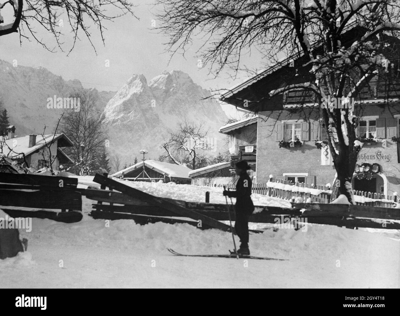 „eine Frau fährt am 10. Februar 1936 durch die olympische Stadt Garmisch. Rechts hat sich das ''Landhaus Berg'' mit einer olympischen Fahne und Kränzen geschmückt, die das olympische Symbol bilden. Im Hintergrund sieht man die Wettersteingebirge mit Höllentalspitzen, Manndl und Waxenstein. [Automatisierte Übersetzung]' Stockfoto