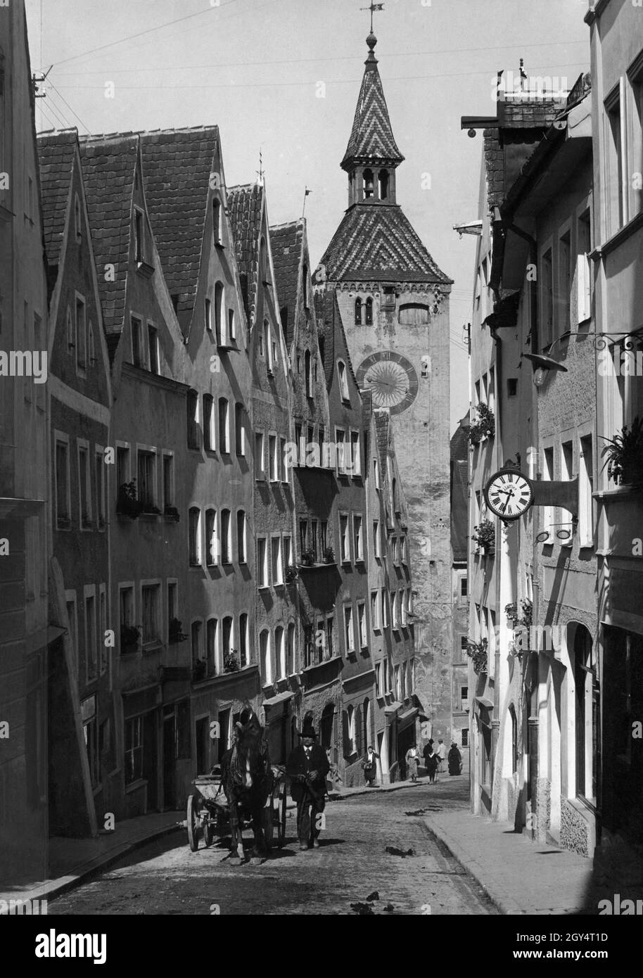 Ein Pferd kämpft die Alte Bergstraße in Landsberg am Lech hinauf. Der Kutscher, gekleidet in feine traditionelle Tracht, geht entlang. Auf der rechten Seite befindet sich das Geschäft eines Optikers. Am unteren Ende der Straße führt der Weg durch den Schmalzturm in den ältesten Teil der Stadt. Undatierte Aufnahme, wahrscheinlich um 1935 aufgenommen. [Automatisierte Übersetzung] Stockfoto