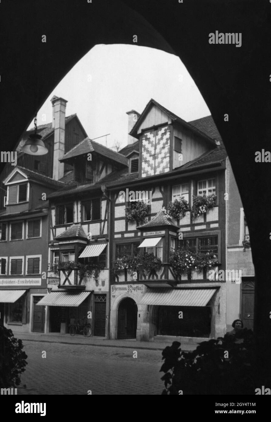 'Das Bild wurde unter einer Arkade des Gasthauses zum Sünfzen aufgenommen und zeigt alte Fachwerkhäuser in der Maximilianstraße in Lindau. Über dem Eingang zum Haus am alten Markt Nr. 4 (Mitte des Bildes) steht ''Hermann Rupflins''. Links davon befindet sich im Haus Nr. 6 ein Friseur. Ganz links im Haus Nr. 8 befand sich im Erdgeschoss das ehemalige Schweizer Getreidehandhaus, die ''Feinbäckerei Ernst H[...]''. Foto undatiert, vermutlich um 1925 aufgenommen. [Automatisierte Übersetzung]' Stockfoto