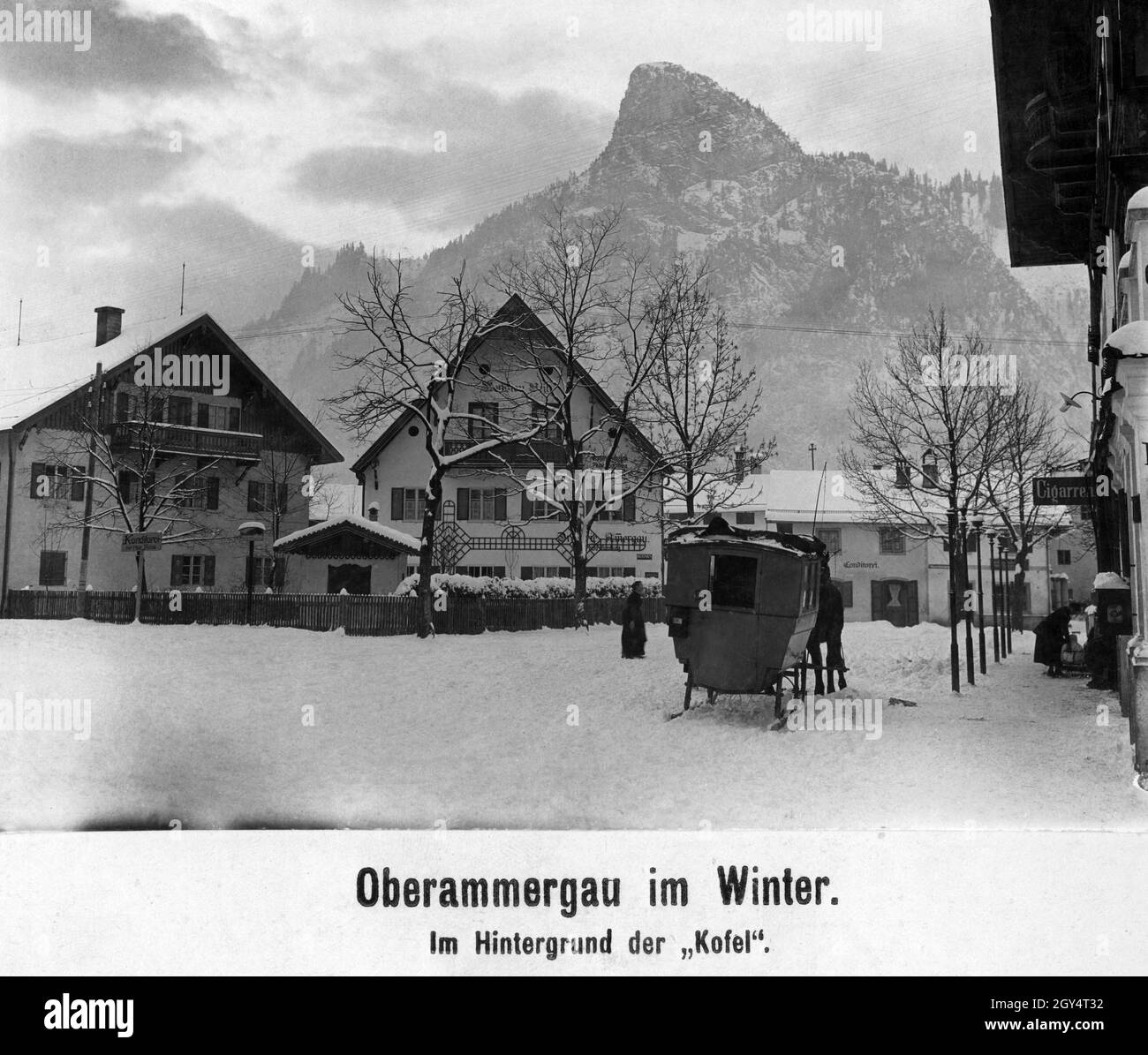 „die Pferde können sich nur mit einem Schlittenwagen durch den Oberammergau, der im Winter 1921 tief im Schnee liegt, durchfahren lassen. Links zeigt ein Schild nach links in die Schnitzlergasse zur Konditorei von Max Hesse. Im Haus in der Bildmitte befindet sich die Drogerie Ammergau mit einer Foto-Handlung, die Kodaks verkauft, und die Pension weil. Rechts auf dem Bürgersteig der Dorfstraße beugt sich eine Frau vor dem Eingang zu einem Zigarrenladen. Der Kofel ist im Hintergrund zu sehen. [Automatisierte Übersetzung]' Stockfoto