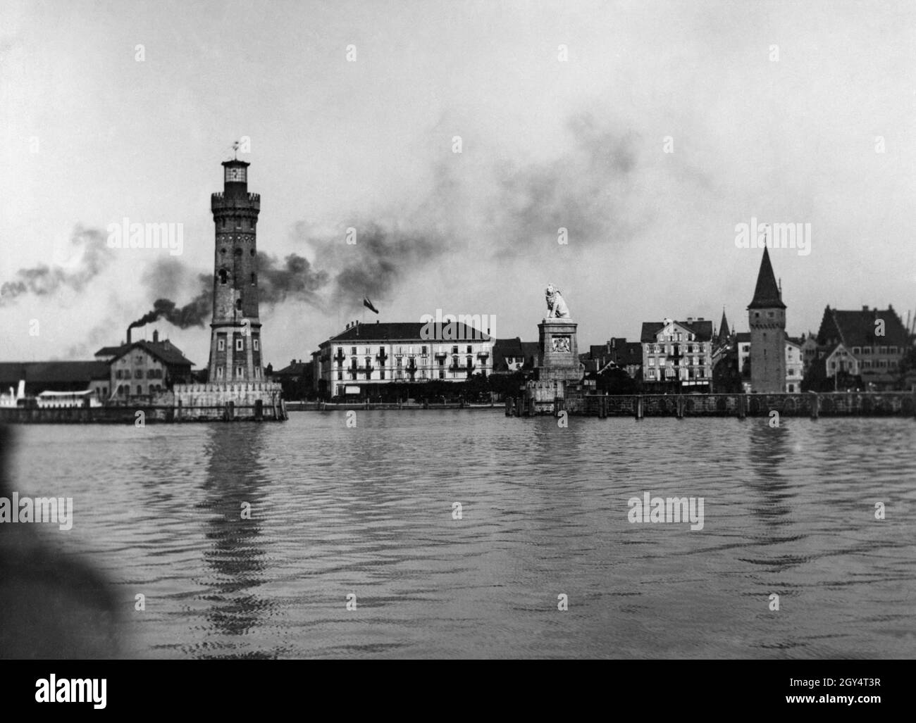 Das Bild zeigt die Hafeneinfahrt zum Hafen von Lindau im Bodensee, die vom Neuen Leuchtturm und dem Bayerischen Löwen begrenzt wird. Weiter rechts befindet sich der Mangturm. Ganz links raucht auf dem Bahnhofsgelände Rauchschwaden. Das Gebäude in der Mitte des Bildes ist das Hotel Bayerischer Hof, weiter rechts ist das Hotel Reutemann zu sehen. Undatierte Aufnahme aus dem Kunstverlag von Max Stuffler aus München, vermutlich um 1910 aufgenommen. [Automatisierte Übersetzung] Stockfoto