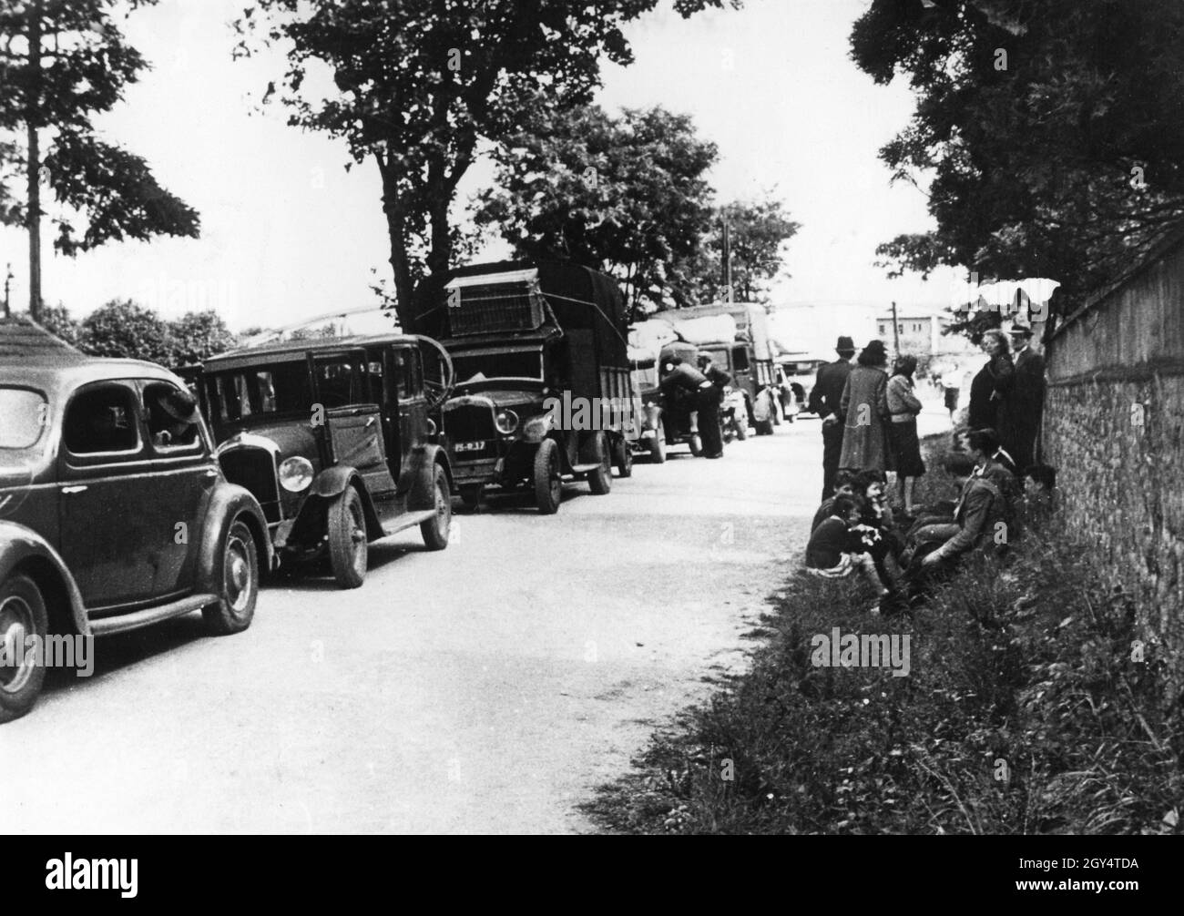 Auf einer Landstraße in Frankreich gibt es Fahrzeuge französischer Flüchtlinge. Am Straßenrand warten die Menschen im Schatten einer Mauer. [Automatisierte Übersetzung] Stockfoto