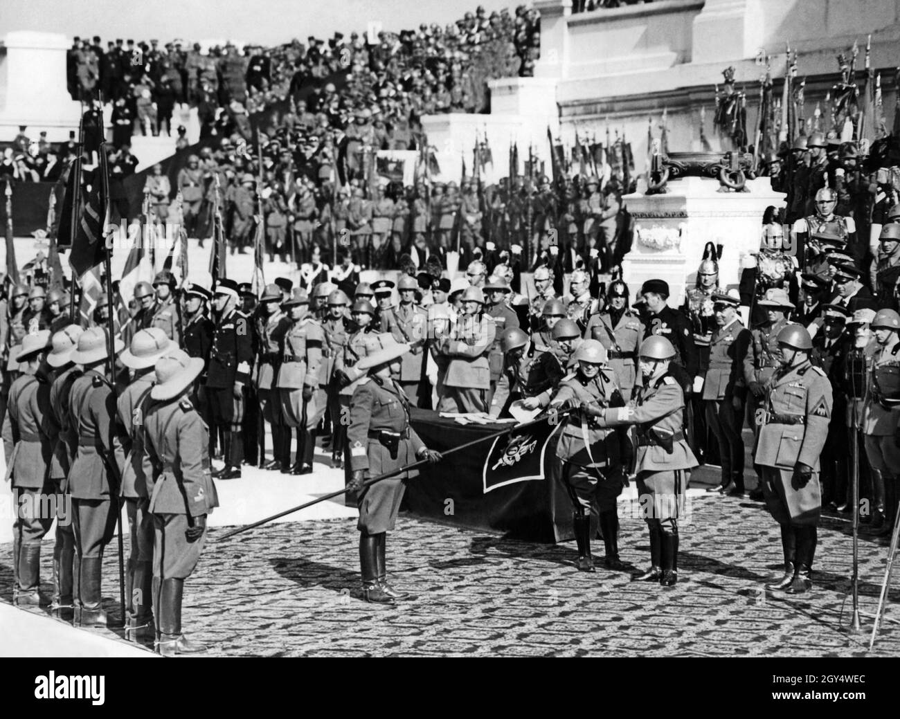 "Am 9. Mai 1937, dem Jahrestag nach der Gründung des ''Italienischen Reiches'', fand in Rom eine große Parade statt, um zu feiern. Am "Altar des Vaterlandes" auf dem Vittoriano in Rom schmückte Viktor Emanuel III., König von Italien (rechts, mit ausgestreckten Armen), die Flaggen, die am Angriffskrieg und der Eroberung Abessiniens teilgenommen hatten. Ein Offizier der Kolonialtruppen hält dem König eine Flagge. Rechts vom König befindet sich Benito Mussolini. Rechts hinter ihm befindet sich General Emilio de Bono (mit weißem Bart). [Automatisierte Übersetzung]' Stockfoto