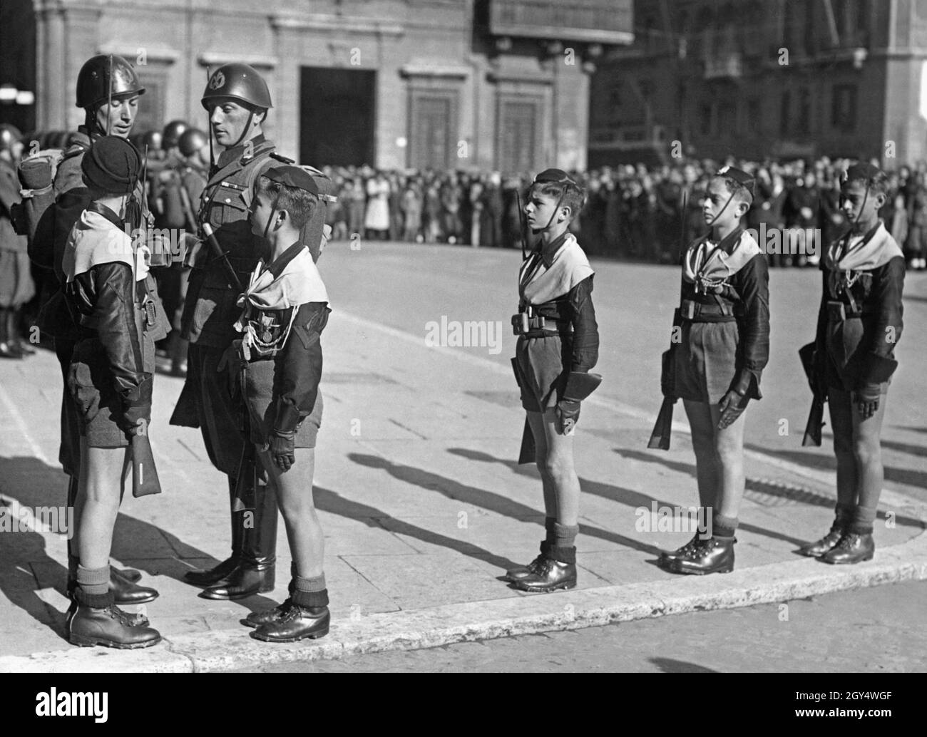 Am Sonntag, dem 29. Oktober 1939, jährte sich die Gründung der Gioventu Italyana del Littorio (GIL) zum zweiten Mal. Der Balilla wurde ebenfalls dem Dachverband untergeordnet. Das Bild zeigt eine Gruppe von Balilla-Jungen, die am Eingang des Palazzo Venezia auf der Piazza Venezia, Rom, Wache stehen, am Tag der Feier. Eine große Menschenmenge beobachtet sie bei der Durchführung ihres Zeremoniells. Zwei italienische Soldaten stehen neben den Jungen. Im Hintergrund sieht man den Palazzo Bonaparte. [Automatisierte Übersetzung] Stockfoto