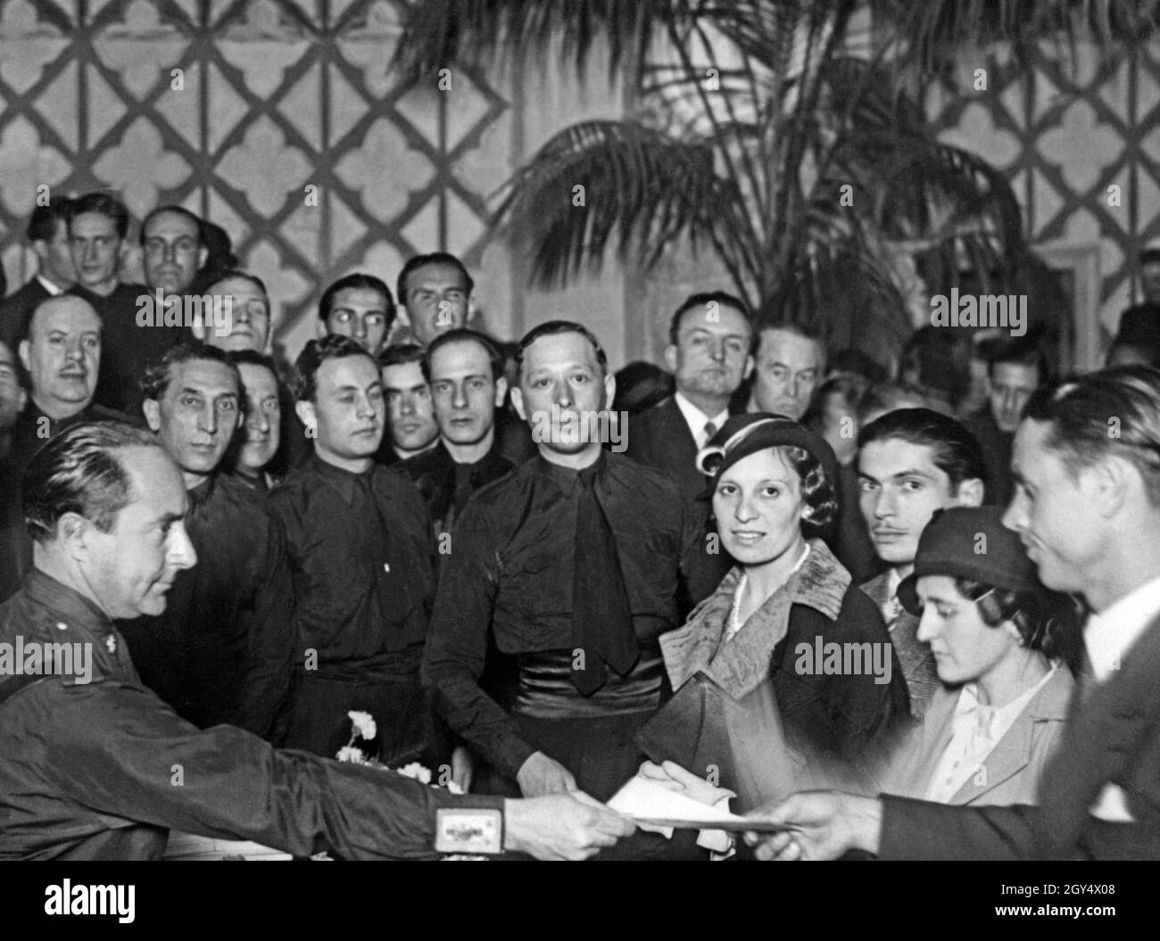 Achille Starace (sitzend, links), Parteisekretär der Partito Nazionale Fascista, überreicht Bräute und Bräutigam im Namen des faschistischen Regimes und Regierungschef Mussolini Hochzeitsgeschenke. Am 30. Oktober 1933 heirateten 2600 Bräute und Bräutigam bei einer großen italienischen faschistischen Veranstaltung in Rom. Rund um den Tisch sind zahlreiche schwarze Hemden. [Automatisierte Übersetzung] Stockfoto