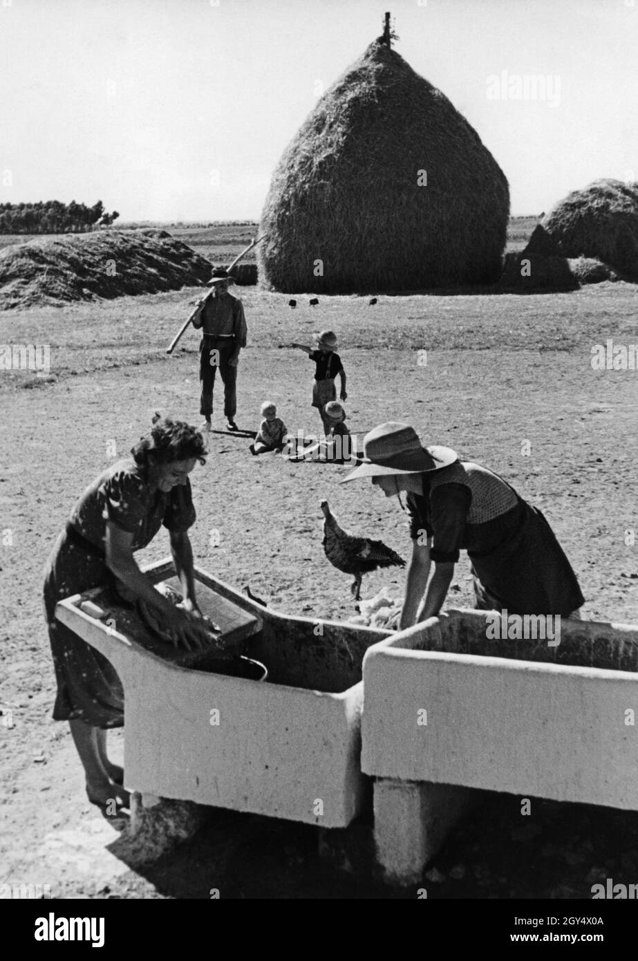 Eine italienische Bauernfamilie in der Kolonie von italienischem Libyen während ihres Arbeitstages. Die Frauen waschen in einem großen Trog Kleidung, im Hintergrund sitzen drei Kinder mit ihrem Vater, der von der Arbeit auf den Feldern mit einer Pitchfork kommt. Im Hintergrund stapelt sich getrocknetes Heu in einem großen Haufen. Undatierte Fotografie, wahrscheinlich in den 1930er Jahren aufgenommen. [Automatisierte Übersetzung] Stockfoto