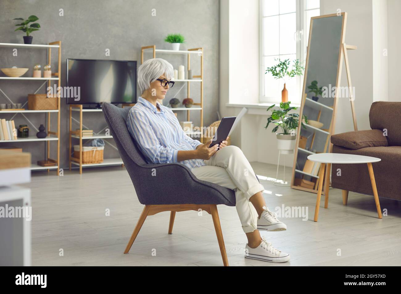 Entspannte ältere Dame, die zu Hause in ihrem Sessel sitzt und ein eBook auf ihrem Tablet liest Stockfoto