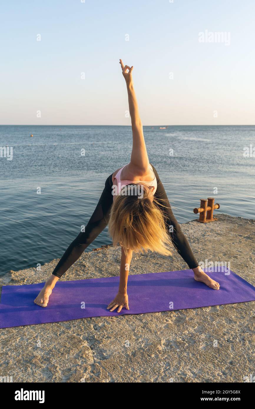 Eine Frau führt die parivrtta padottanasana Verdrehung Übung durch, stehend auf dem Pier, Training an einem sonnigen Sommerabend Stockfoto