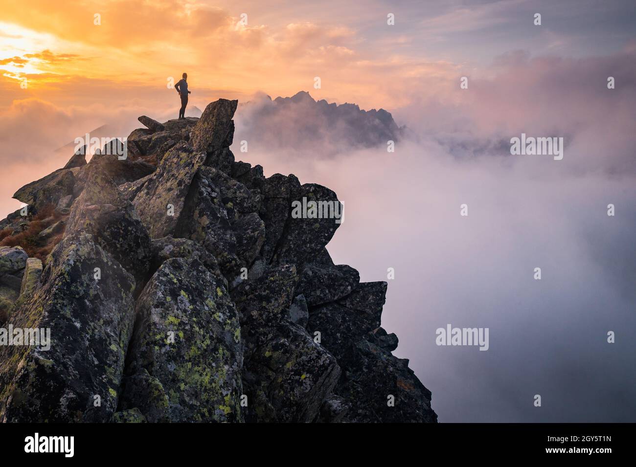 Der Mensch steht allein auf dem Gipfel des Felsens. Wanderer beobachten die Herbstsonne am Horizont. Schöner Moment das Wunder der Natur. Bunter Nebel im Tal. Mann h Stockfoto
