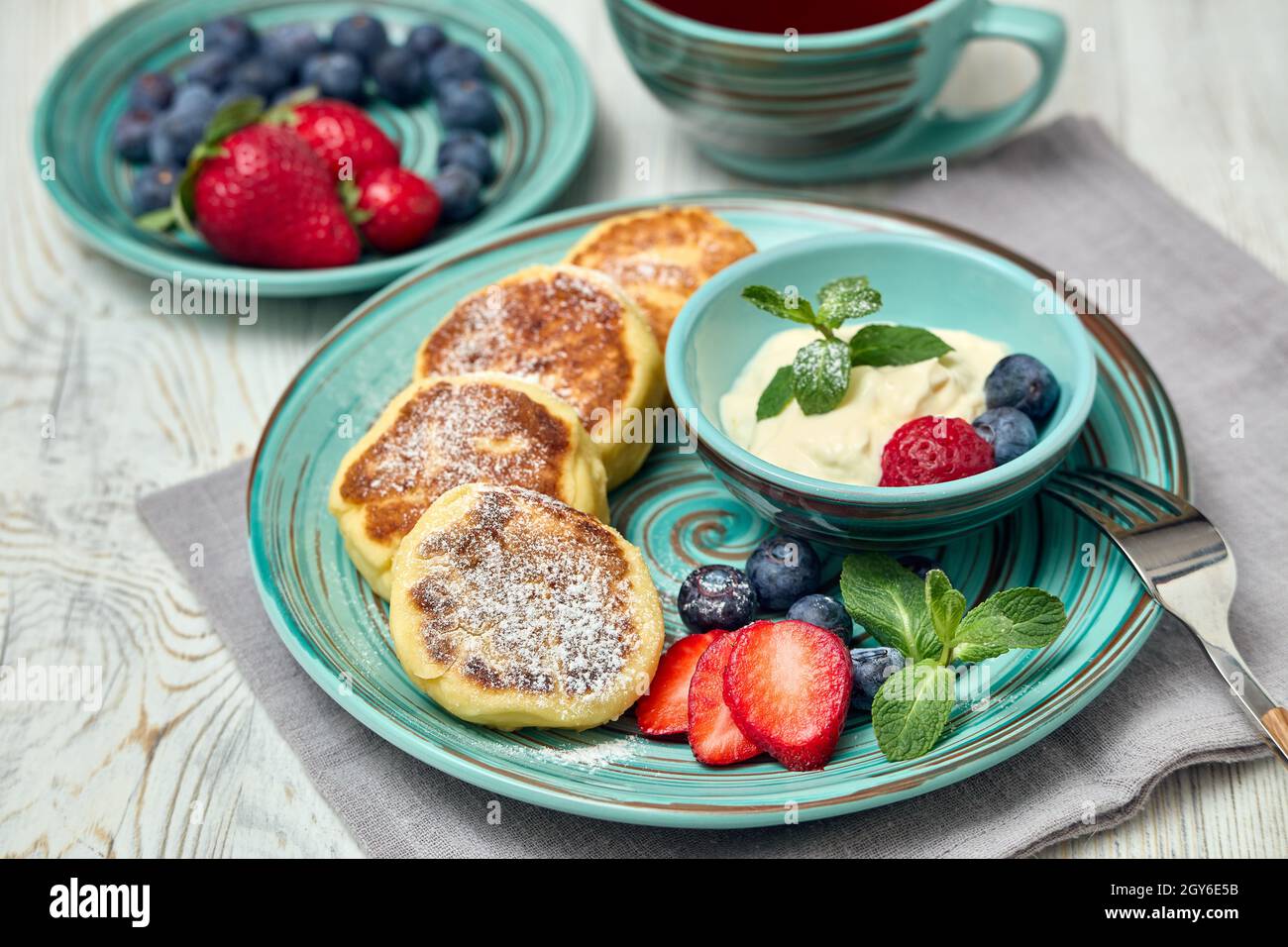 Russische Hüttenkäse-Pfannkuchen (Syrniki) mit Beeren und saurer Creme, Tasse mit Früchtetee auf hellem Holztisch mit Sonnenlicht vom Fenster. Gesunde Farbtonfarbe Stockfoto