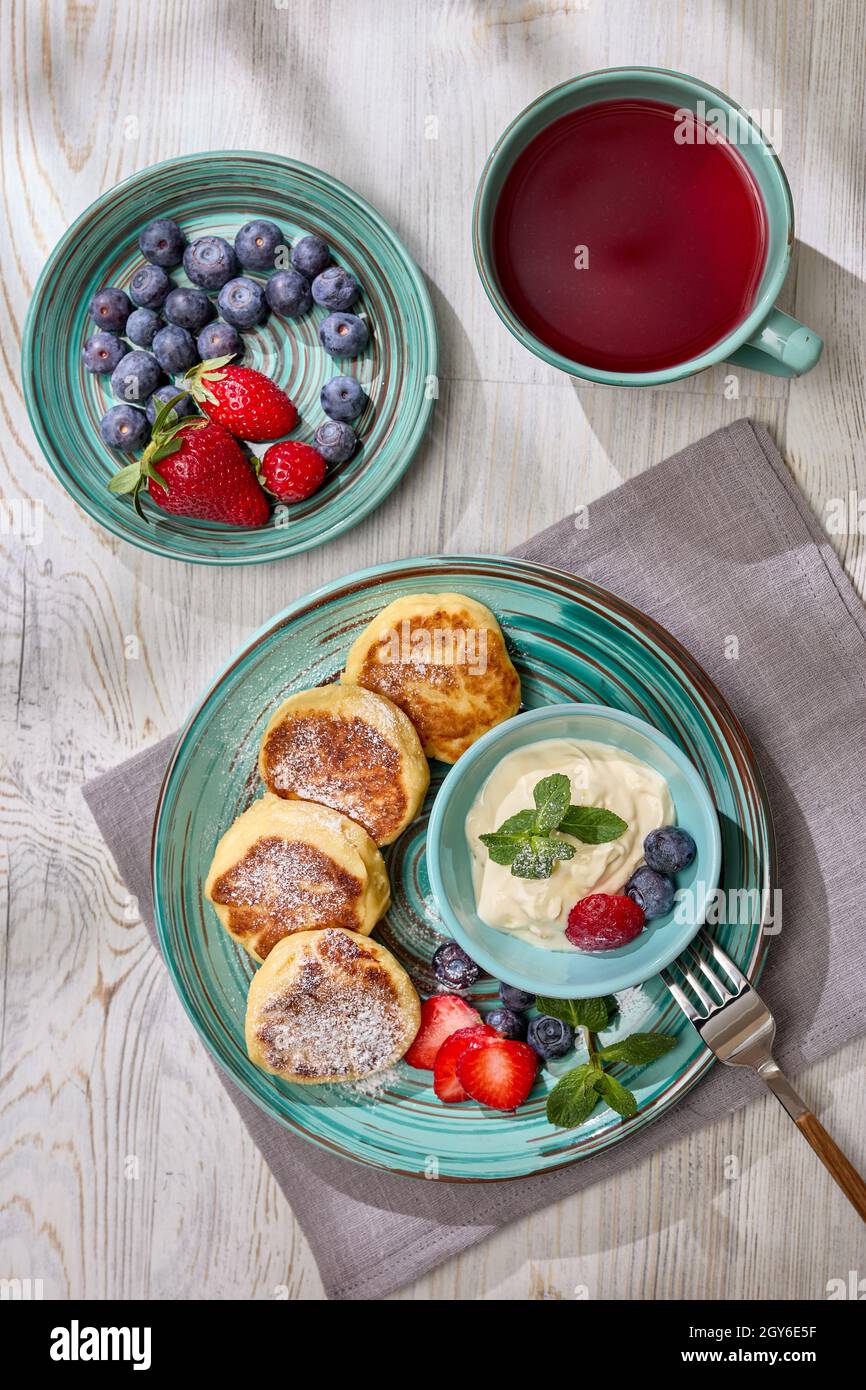 Russische Hüttenkäse-Pfannkuchen (Syrniki) mit Beeren und saurer Creme, Tasse mit Früchtetee auf hellem Holztisch mit Sonnenlicht vom Fenster. Draufsicht. H Stockfoto