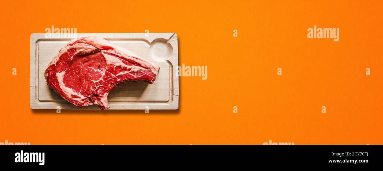 Rohes Rindfleisch Prime Rib und Holz Schneidebrett isoliert auf orangefarbenem Hintergrund. Draufsicht. Horizontales Banner Stockfoto