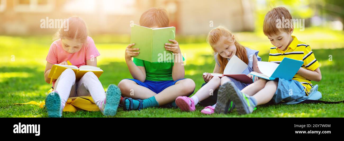 Kinder mit Rucksäcken sitzen im Park auf dem Gras in der Nähe der Schule. Schüler mit Büchern und Rucksäcken im Freien. Stockfoto