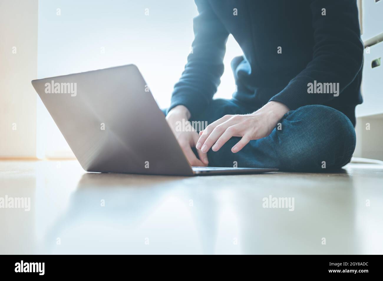 Junger Mann mit einem Laptop arbeitet auf dem Holzboden, Licht aus dem Fenster kommt Stockfoto