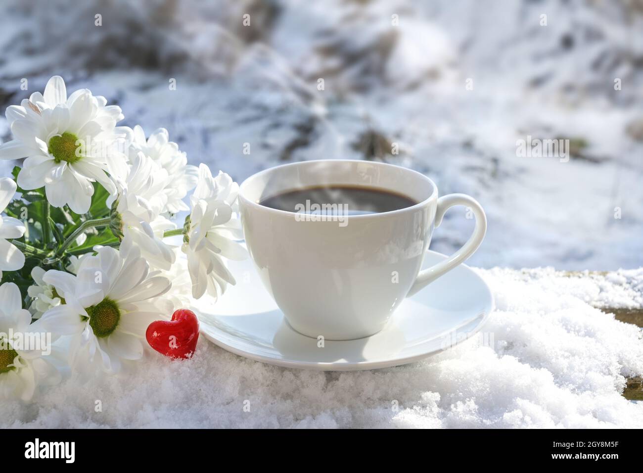 Frühstück im Freien im Winter mit Liebe, dampfende Kaffeetasse, weiße Blumen und ein rotes Glas Herz auf einem schneebedeckten Gartentisch an einem sonnigen Tag im n Stockfoto