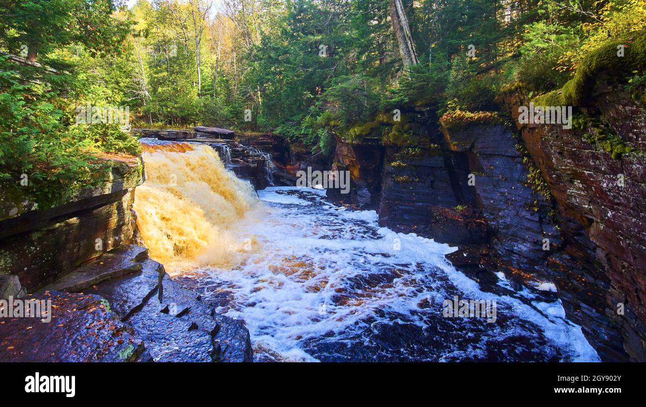 Blauer Fluss mit goldenem und braunem Wasserfall in einer kleinen Schlucht mit Wald auf beiden Seiten Stockfoto