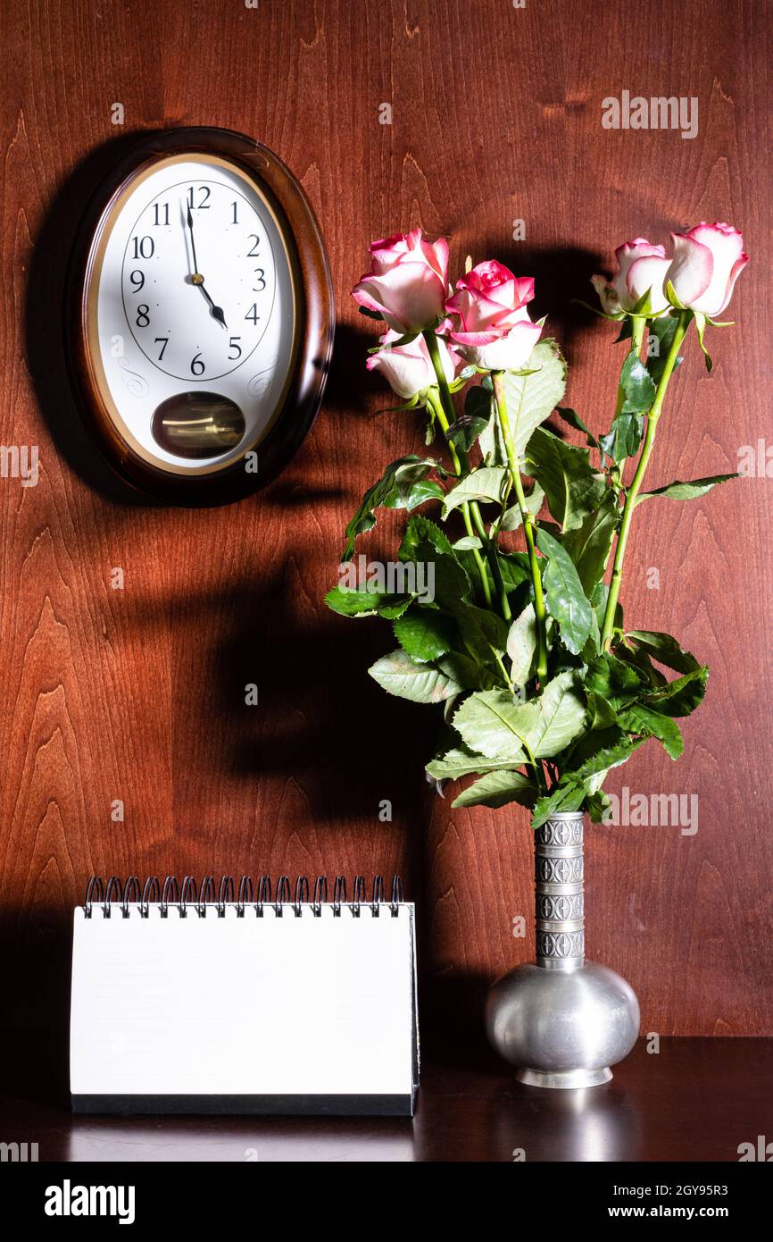 Wanduhr, leerer Kalender und rosa Rosen in Zinnkrug auf dunkelbraunem Holzhintergrund Stockfoto