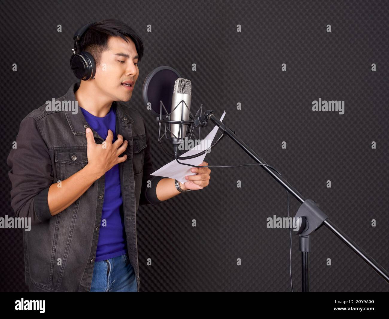 Musiker, die Musik im professionellen Aufnahmestudio produzieren. Ein junger asiatischer Mann mit Kopfhörern singt, während er den Text in der Hand vor dem schwarzen so hält Stockfoto