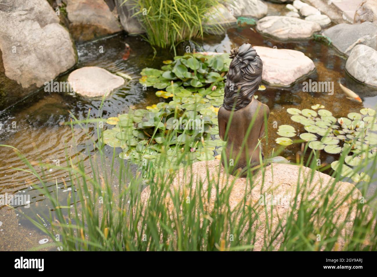 Teich im Park. Künstliche Landschaft mit Steinen und Wasser. Eine stählerne Meerjungfrau sitzt auf einem Felsen zwischen Veilchen. Stockfoto
