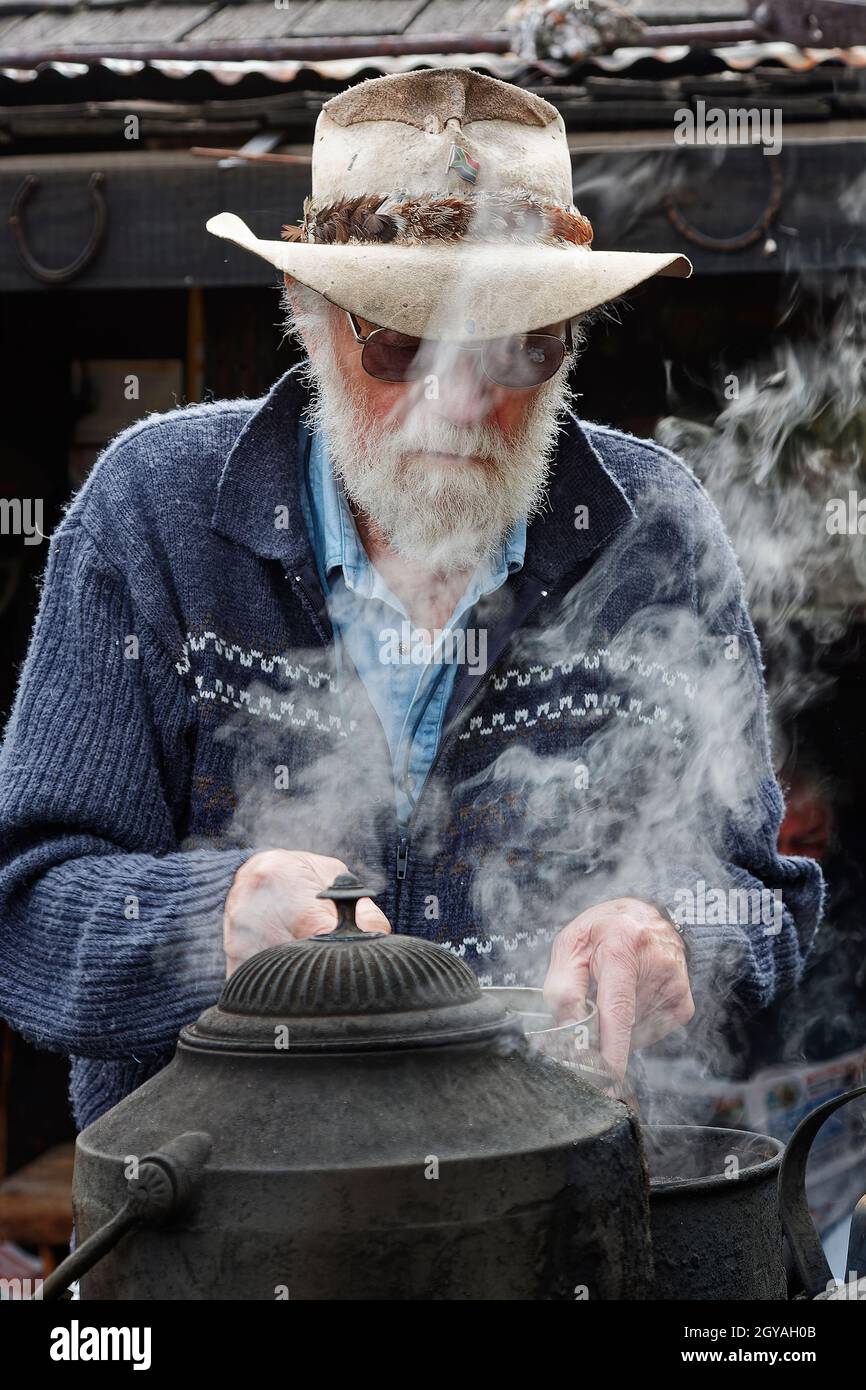 billy-Tee zubereiten; älterer Mann, Hut, Pullover, weißer Bart, Dampf, alte Ausrüstung, Erfrischung, Bearded Mining Co.; The Miners' Hut, Reefton; Neuseeland Stockfoto