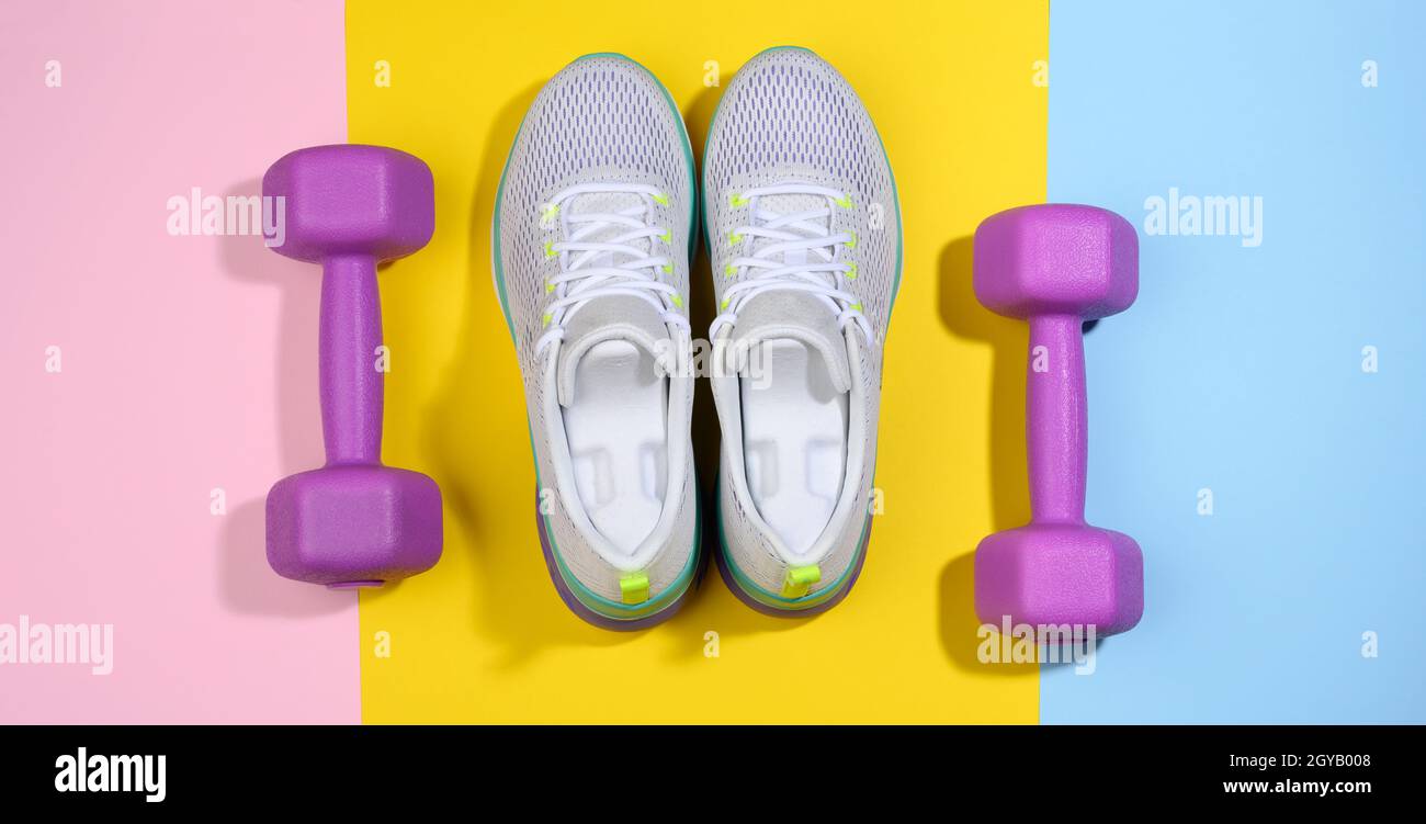 Ein Paar weiße Damen-Sneaker und lila Hanteln auf einem bunten Hintergrund, Sport Stockfoto