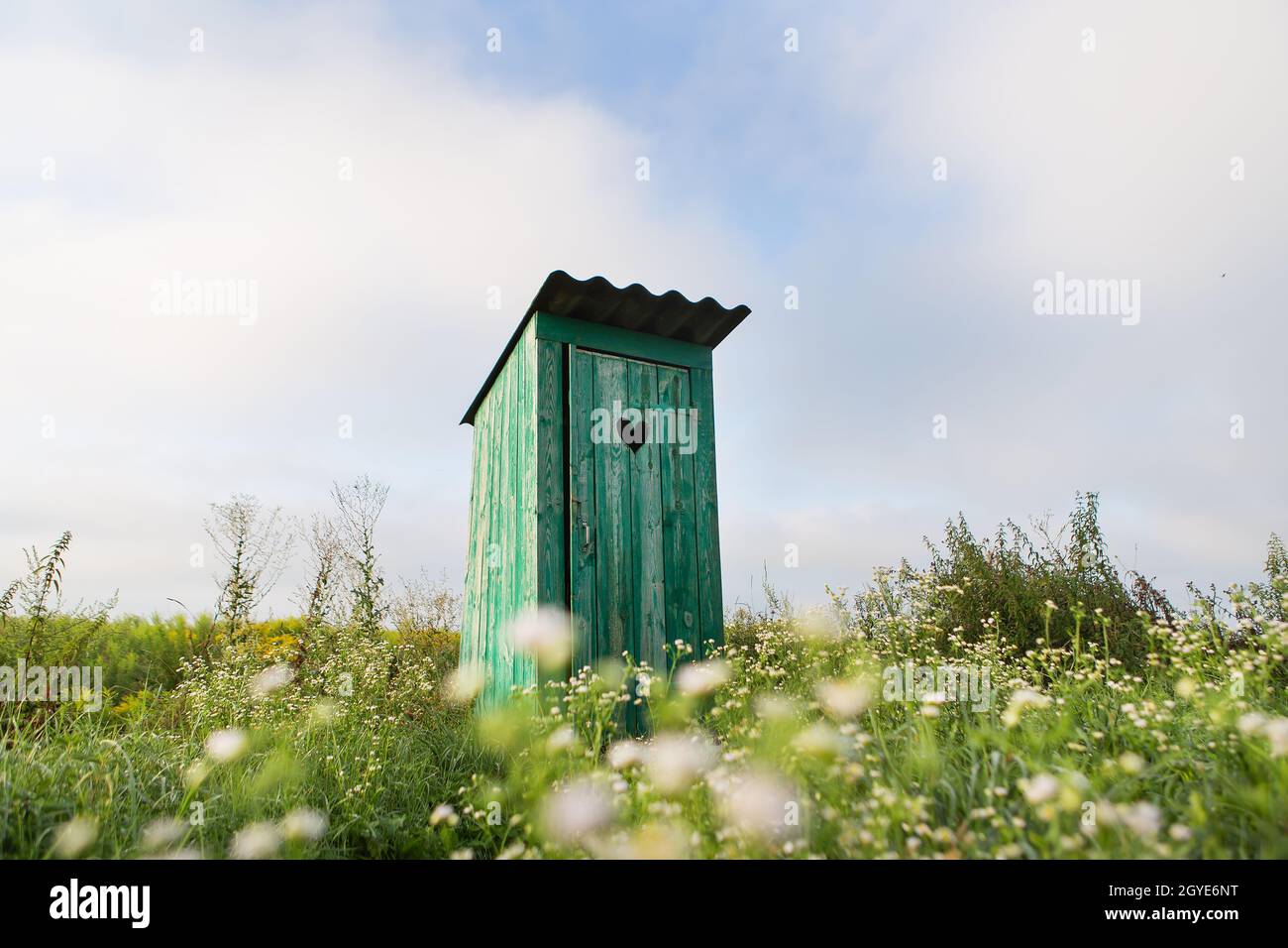 Vintage-Toilette. Eine rustikale grüne Toilette im Freien mit einem Herz, das an der Tür herausgeschnitten wurde. Toilette in einem Blumenfeld Stockfoto